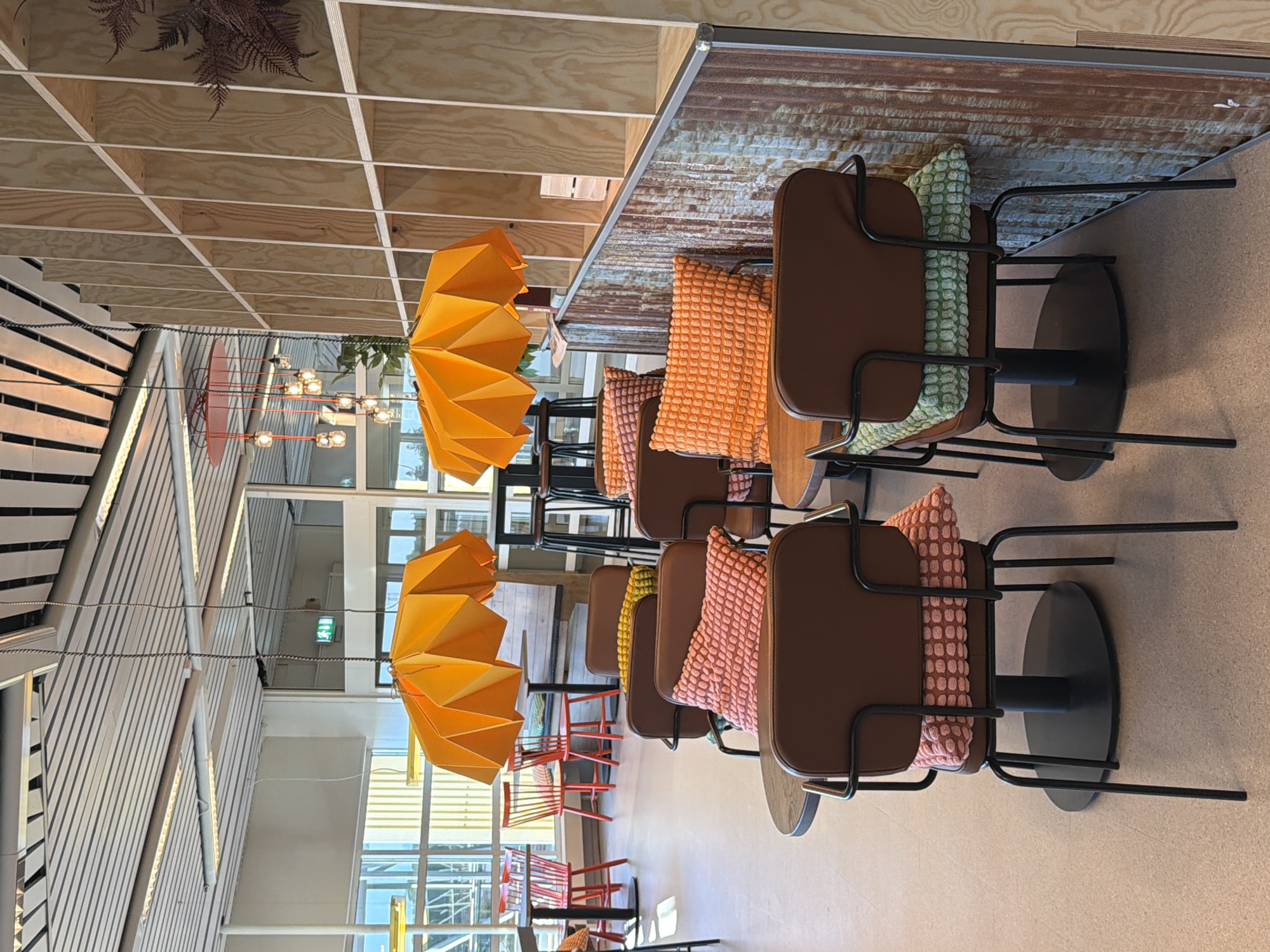 This image depicts the interior of a modern cafe, likely located at Malmö-Sturup Airport, Sweden, judging by the presence of a green emergency exit sign. The cafe features a warm and inviting ambiance with natural wood accents and pops of color.
In the foreground, several chairs with brown leather seats and backs are arranged around small, round tables. Each chair is adorned with a decorative cushion: some are pink with a textured pattern, while others are bright orange or light green. The chairs have slender black metal frames and legs, contributing to a contemporary aesthetic.
Dominating the mid-ground are two large, origami-like lampshades in a vibrant orange hue, suspended from the ceiling. These lamps cast a warm glow, complementing the overall cozy atmosphere. Behind the chairs, a wall paneling made of light-colored wood is visible, featuring a grid-like pattern of shelves. A section of this wall has a textured, rustic finish with blue and brown tones, adding visual interest.
In the background, through large windows, glimpses of the outdoors and other parts of the airport terminal can be seen. Red chairs and tables are visible further in the distance, suggesting other seating areas within the cafe or adjacent spaces. The ceiling is modern, with integrated lighting and slatted panels. The floor is a neutral-toned, speckled material, common in public spaces. No people are present in the image, giving the scene a still, observational quality.