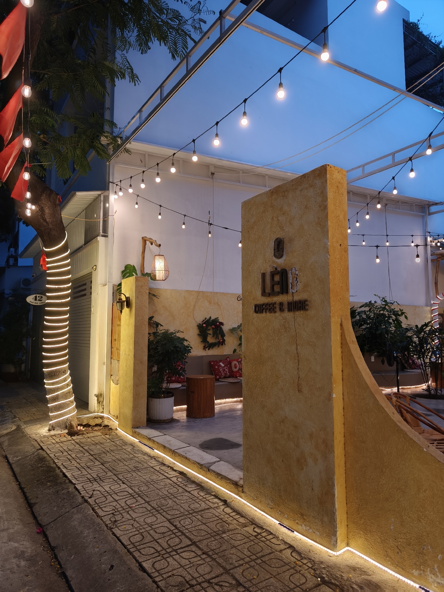 This image depicts the exterior of a coffee shop called "LÊM COFFEE & MORE" at dusk in Bac Nha Trang Ward, Vietnam. The shop features a warm and inviting ambiance, enhanced by strings of illuminated bulbs and decorative lights.
The scene is set with a prominent, textured yellow wall bearing the coffee shop's name. To the left, a tree wrapped in a spiral of bright white lights stands beside a building with a "42" address marker. A sidewalk paved with patterned tiles leads towards the entrance, bordered by more subtle strip lighting.
Red flags hang from the tree, adding a touch of color against the darkening sky and lush green foliage. Inside the covered seating area, comfortable seating with red cushions is visible, along with a decorative wreath and potted plants. A wooden stool sits in the foreground of the seating area. The overall mood is relaxed and cozy, suggesting a pleasant evening gathering place. The time of day is clearly evening or twilight, with artificial lights being the primary source of illumination, casting a warm glow on the surroundings.