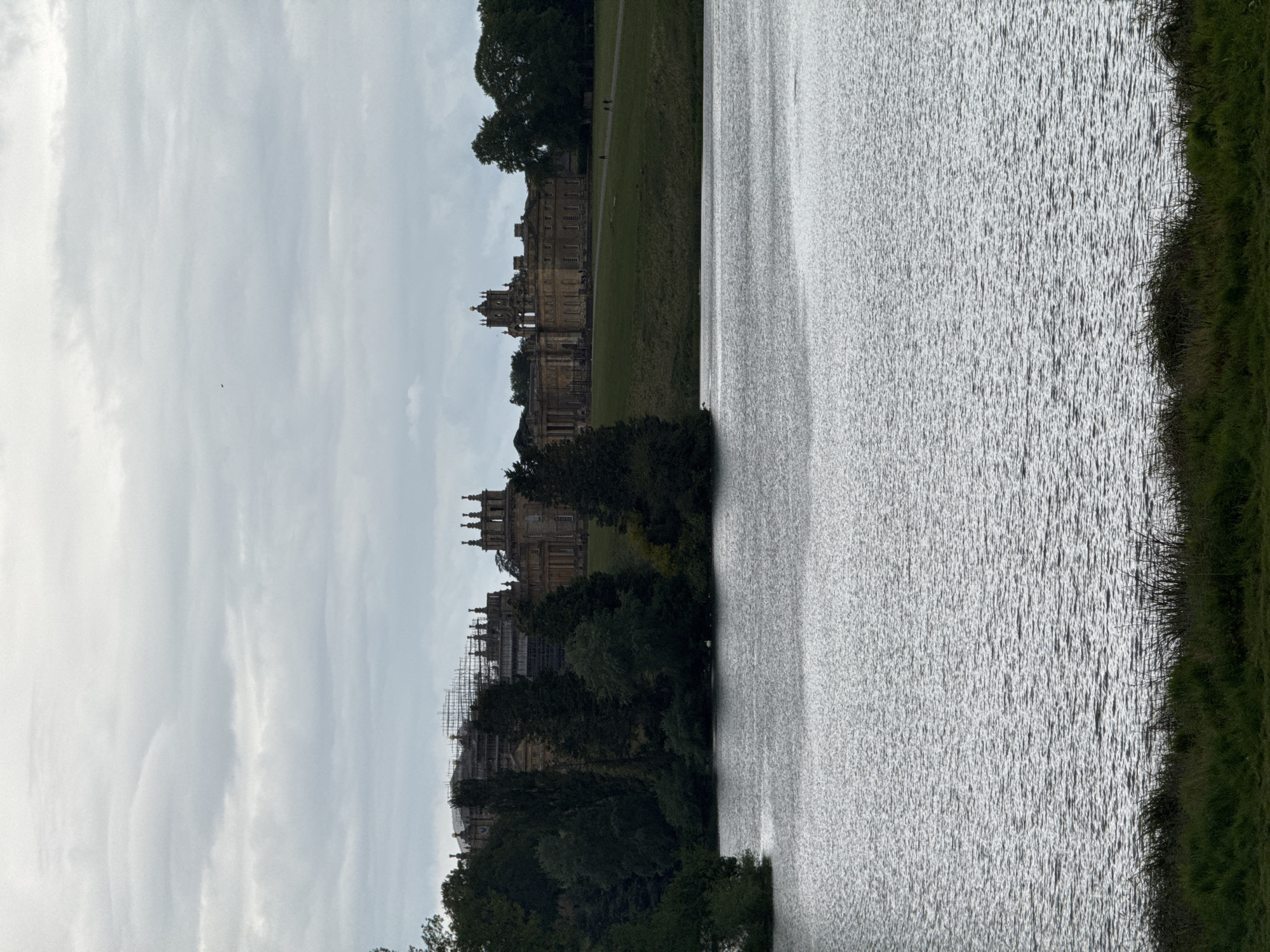 The photograph depicts Blenheim Palace, located in Woodstock, West Oxfordshire, United Kingdom, as viewed from across a body of water. The palace, an imposing structure, is partially obscured by trees in the foreground. Scaffolding is visible on a section of the palace, indicating ongoing construction or restoration work. The water surface is textured, reflecting the overcast sky. The far shoreline consists of grass. The sky is cloudy. Two small figures are walking on the grass next to the palace.