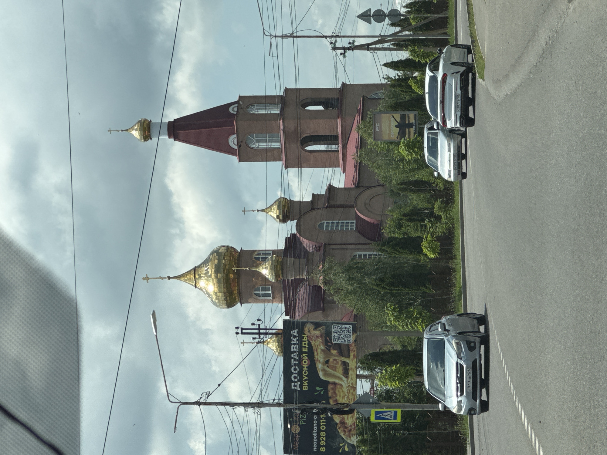 The image captures a street view in Mikhaylovsk, Russia, featuring a prominent Orthodox church with golden domes and a red spire. In the foreground, a silver Subaru Impreza is driving on a paved road, with its license plate reading "E717HH 126". Behind it, two other cars are visible: a white sedan on the left and a white Kia on the right. A billboard advertising food delivery is mounted on a pole to the left of the church. The sky is overcast with clouds, and power lines crisscross the upper portion of the image. Lush green trees line the side of the road and surround the church.