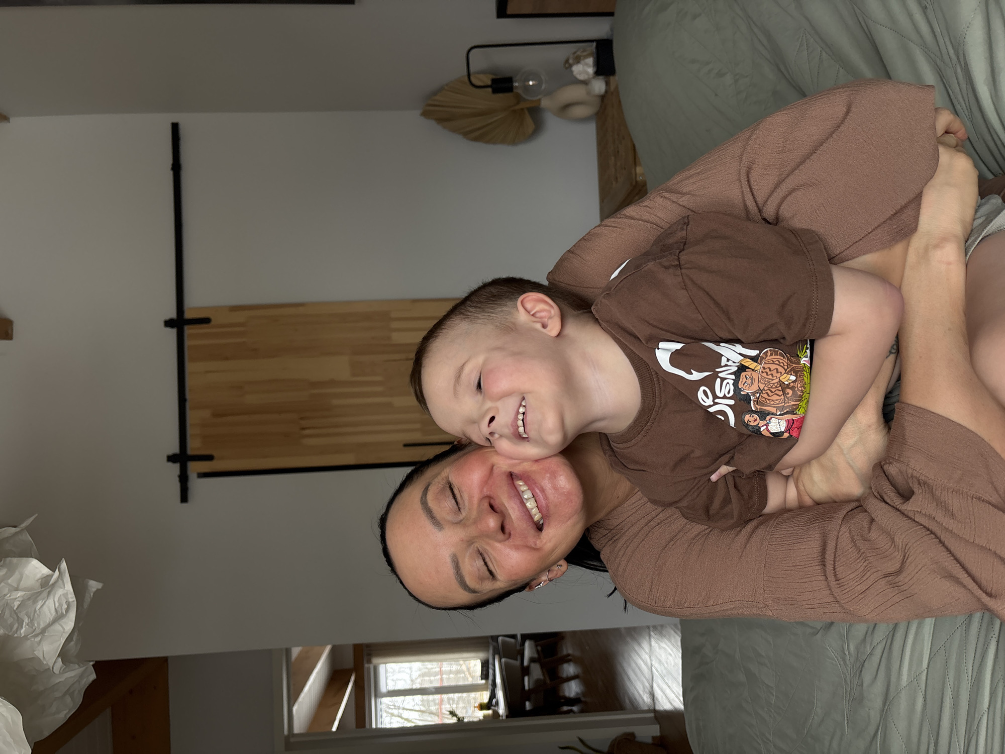 A woman and a child are embracing indoors. The woman is wearing a brown outfit, and the child is wearing a brown T-shirt. They are both smiling. A bed with a green comforter is visible in the background. The room has a wooden sliding door and a window. The location is potentially Chaikovsky, Russia, based on contextual cues not evident in the image itself.
