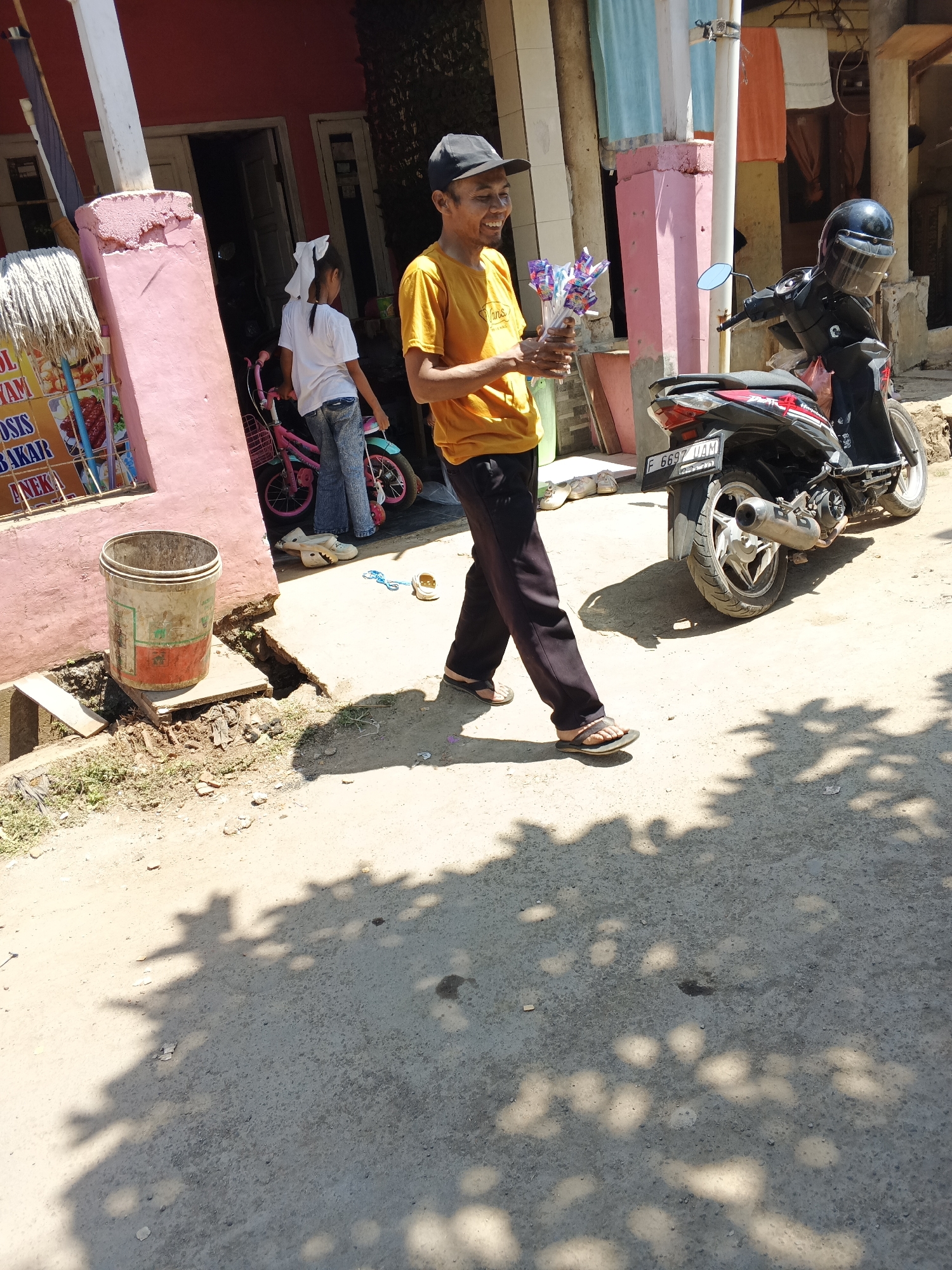In Sukaharja, Indonesia, a young man wearing an orange t-shirt and a black cap walks on a dirt road, holding a bunch of colorful candies. He appears to be a street vendor. To his left, a young girl in a white t-shirt and jeans stands beside a pink bicycle. A black motorcycle is parked behind the man. The background features a pink building and some laundry hanging to dry. The ground is dusty and uneven, with shadows from overhead trees dappling the scene.