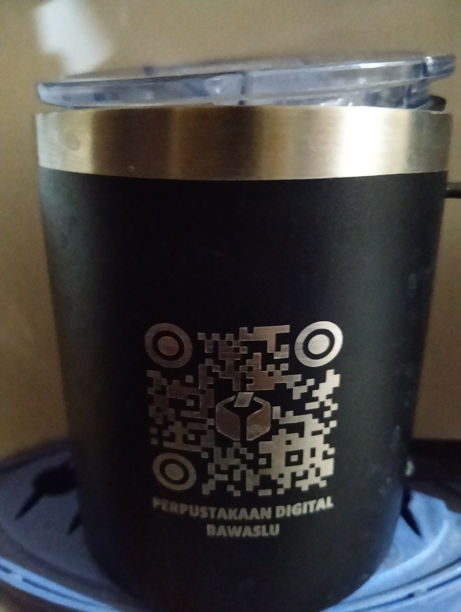 A black insulated tumbler with a metallic band and a clear lid is shown, featuring a QR code and text that reads "PERPUSTAKAAN DIGITAL BAWASLU". The tumbler is placed on a blue surface, possibly a mat or cloth, with a blurred background suggesting an indoor environment. The lighting is moderate, casting some shadows on the tumbler. There are no people or any recognizable landmarks of the city of Payagaboh, Indonesia, visible in the image.