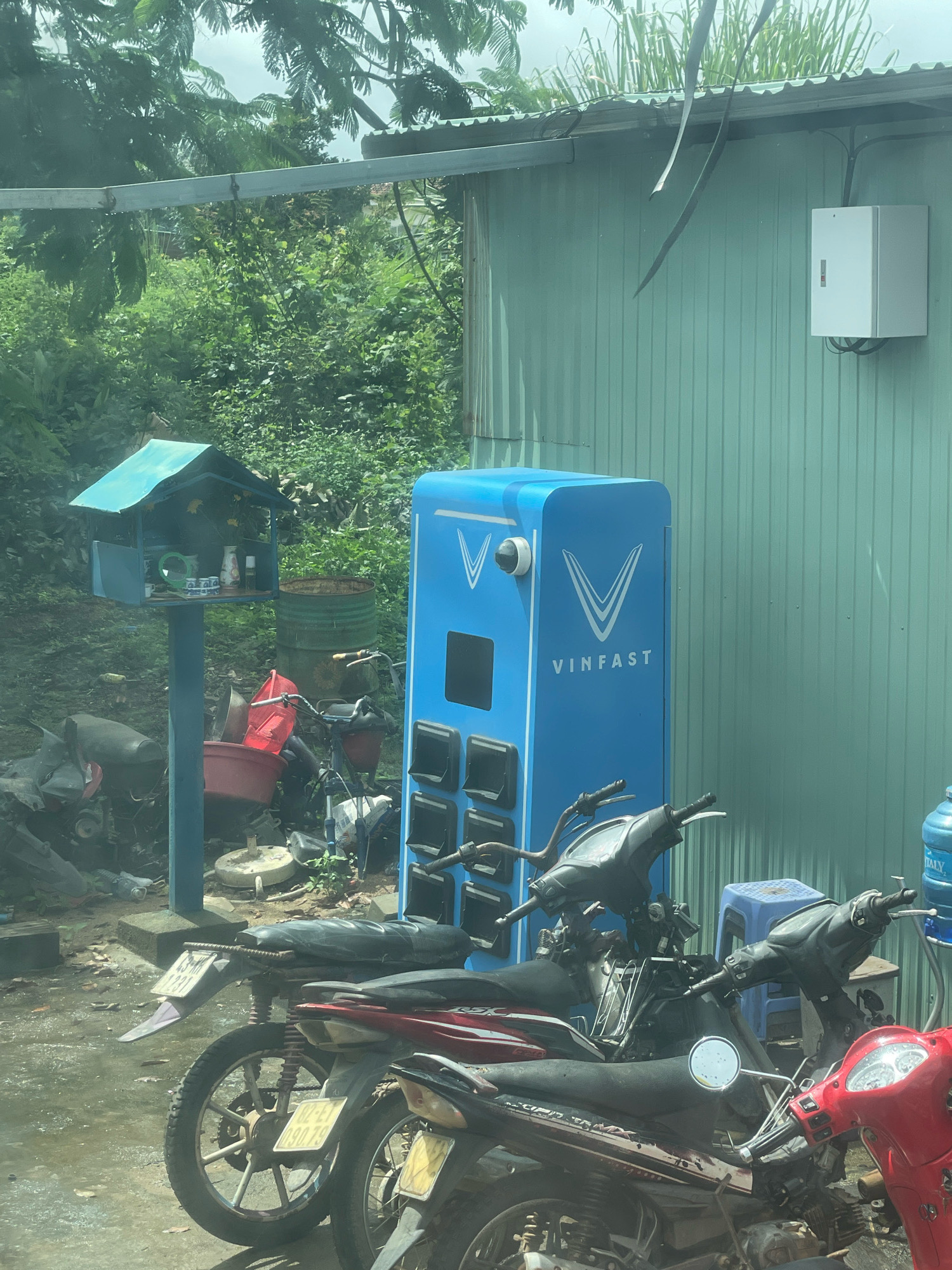 The image depicts an outdoor scene in Pờ Y, Vietnam, showing a VinFast charging station and several parked motorcycles. The VinFast charging station is a tall, bright blue structure with the VinFast logo prominently displayed. It has a camera at the top and a panel with multiple charging slots. To the left of the charging station, there is a small, teal-colored structure resembling a shrine or altar, situated on a blue pole. This shrine contains small decorative items and what appear to be cups and bottles. Behind the shrine, there are various objects including a red plastic bucket, a green barrel, and parts of disassembled motorcycles. A bicycle with a red seat is also visible. In the foreground, several motorcycles are parked, one of which is red, and others are darker in color. The ground is wet and appears to be dirt or gravel. The background features lush green vegetation and a light green corrugated metal building. A white electrical box is mounted on the exterior of this building. The sky is overcast. There are no people visible in the image.