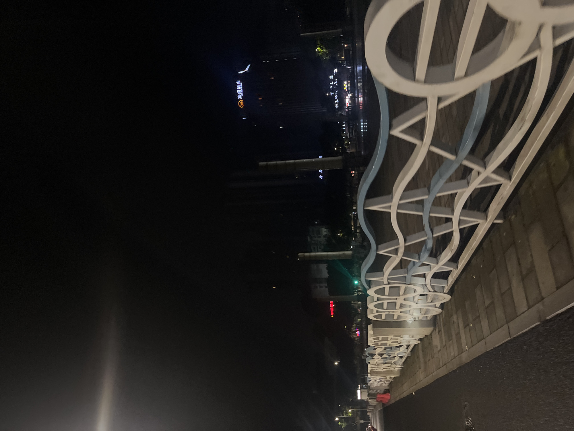The image presents a nighttime urban landscape in Jiulongpo District, China. A paved sidewalk, constructed from rectangular light-colored pavers, extends from the lower-left foreground towards the center-right. Parallel to the sidewalk and immediately to its right, a white and light blue decorative metal fence delineates a boundary. The fence features wavy horizontal top rails, interspersed with circular and triangular motifs.
Beyond the fence, a body of water reflects various artificial lights from the opposite bank. To the left of the sidewalk, an asphalt road surface is visible, with a partial orange traffic cone positioned on its far left edge.
The opposing bank of the water features multiple illuminated structures. A prominent high-rise building on the right displays an orange, illuminated logo and Chinese characters identifying it as "嘉樂酒店" (Jia Le Hotel). Other buildings on this bank exhibit various red and green light sources, including what appears to be a green traffic signal. Several tall, dark cylindrical pillars are vertically prominent within the midground.
The scene is predominantly illuminated by artificial light sources, including streetlights (one producing a significant lens flare in the upper-left) and the lights emanating from buildings. The sky above is dark. No human figures or discernible moving vehicles are present within the frame.