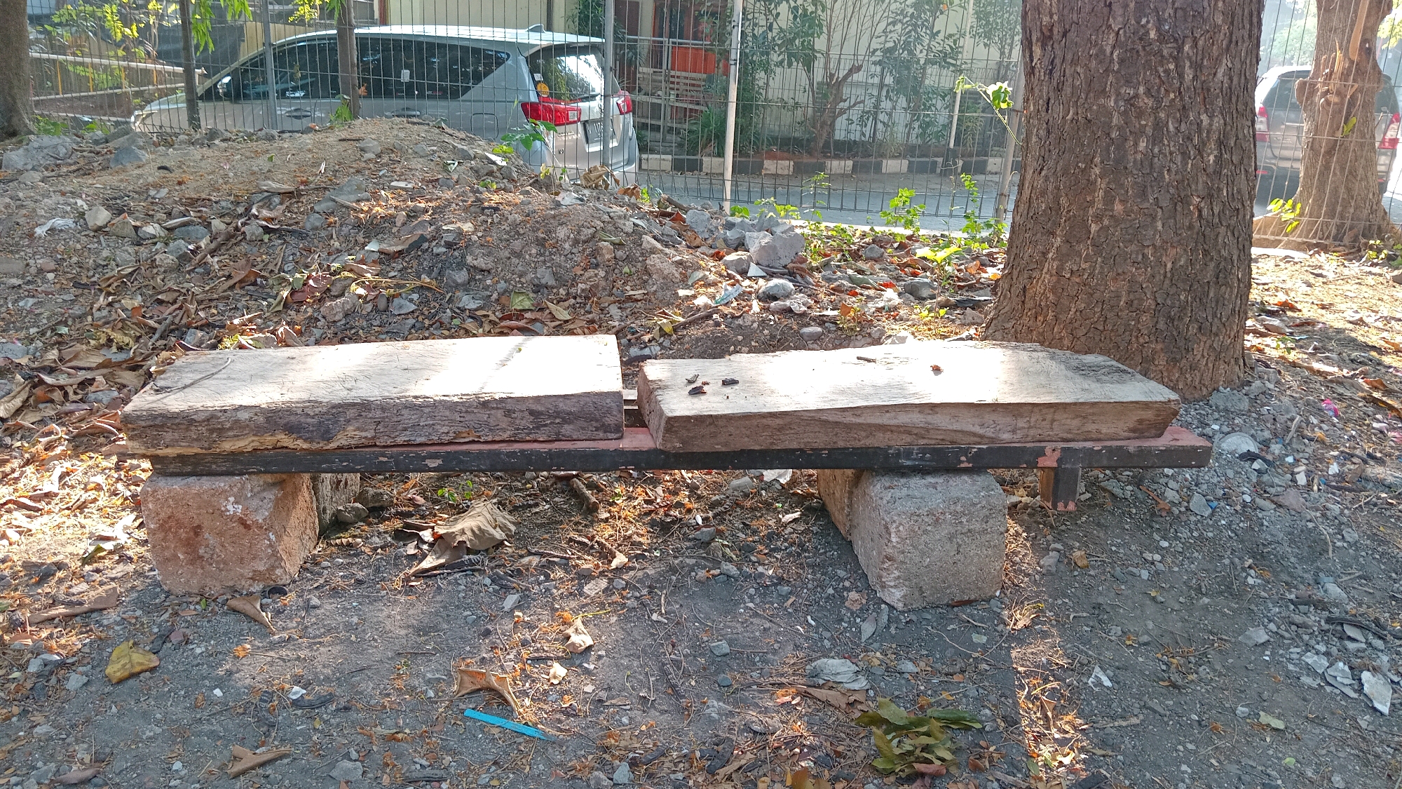In Randuagung, Indonesia, a weathered, rustic bench is situated outdoors. The bench is constructed from two thick, rough-hewn wooden planks resting on a dark metal frame. The frame is supported by two large, irregular concrete blocks. The bench is positioned on a dirt and gravel surface littered with fallen leaves and small stones. In the background, a large tree trunk with textured bark stands prominently to the right. Behind the tree and further back, a silver car is visible through a metal fence, and residential buildings can be seen. The scene is illuminated by sunlight, casting shadows and highlighting the textures of the natural and man-made elements.