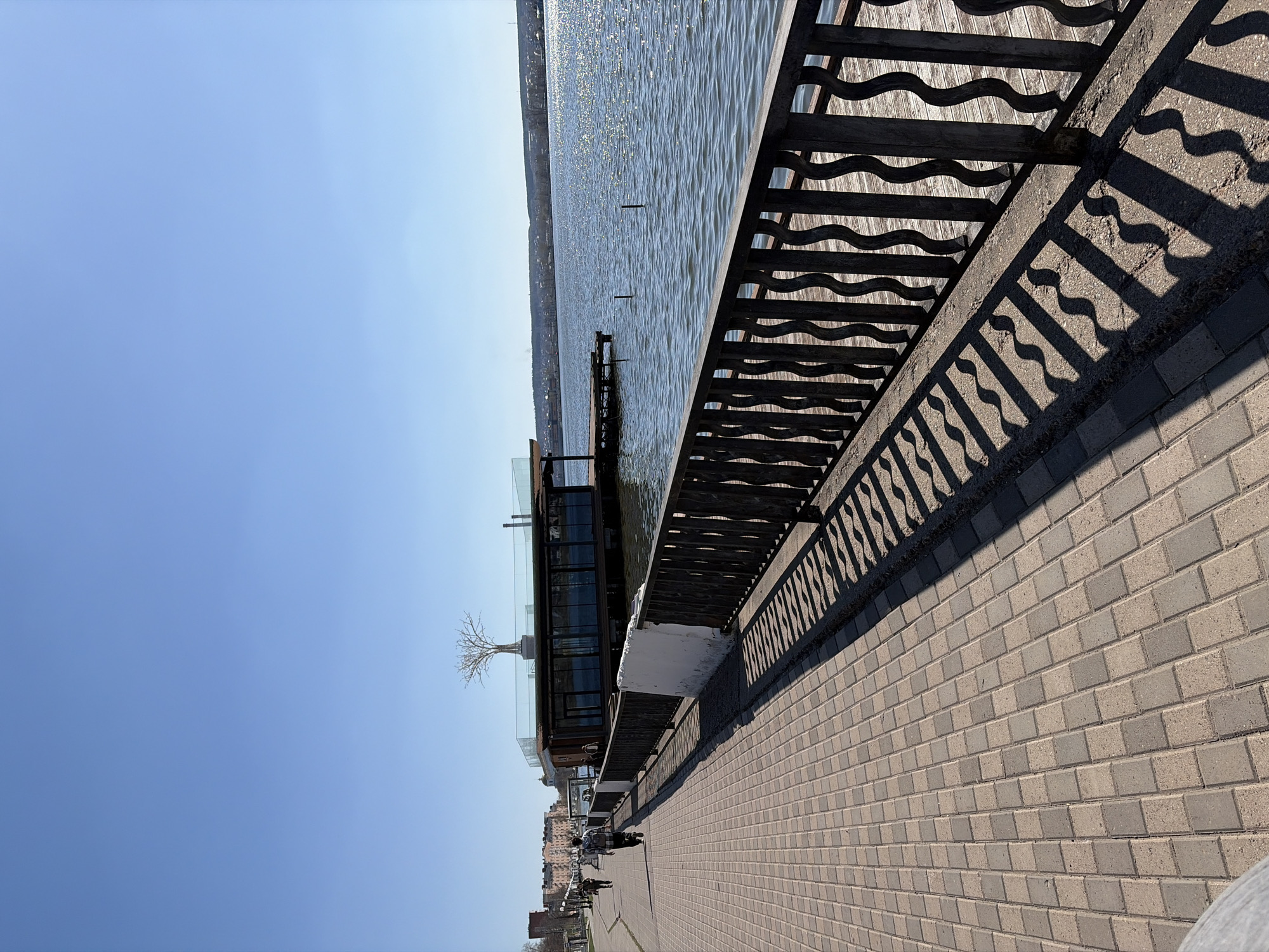 The image depicts a waterfront scene in Votkinsk, Russia, showing a brick-paved walkway bordered by a decorative metal railing. The railing casts distinct shadows on the walkway. To the left of the walkway is a building partially extending over the water, featuring glass panels and a small tree on the roof. Beyond the building is the body of water with slight ripples and light reflecting on the surface. People are visible walking along the walkway in the distance. The sky is a clear, light blue.