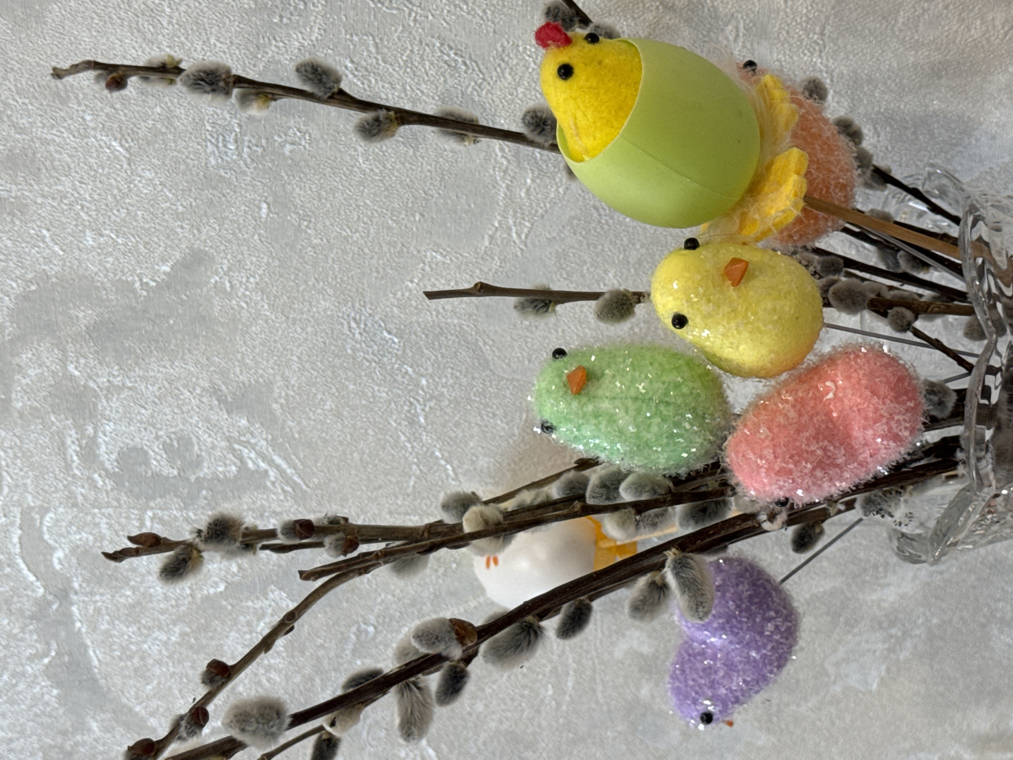 This close-up image displays a festive Easter or spring decoration, primarily featuring pussy willow branches adorned with an assortment of colorful, glittery chick and egg figures. The composition is set against a light gray, textured background, possibly a wall.
On the right side of the frame, several dark brown pussy willow branches, bearing their characteristic soft, fuzzy catkins, are arranged vertically, likely in a clear glass vase which is partially visible on the far right. Scattered among these branches are numerous decorative chicks and egg shapes in vibrant, pastel colors.
At the top, a yellow chick with a small red comb and black eyes emerges from a light green plastic eggshell. Below it, an orange-colored glittery chick with yellow wings is visible. Further down, there are a bright yellow glittery chick with black eyes and an orange beak, a green glittery chick with similar features, and a pink glittery egg-shaped decoration with black eyes. A white chick with an orange beak is subtly nestled among the branches, and at the bottom, a purple glittery chick with black eyes and an orange beak completes the cheerful arrangement.
The background is uniformly light gray with a rough, stucco-like texture, suggesting an indoor setting. The overall scene evokes a sense of spring, renewal, and celebration associated with Easter. No text is visible in the image.