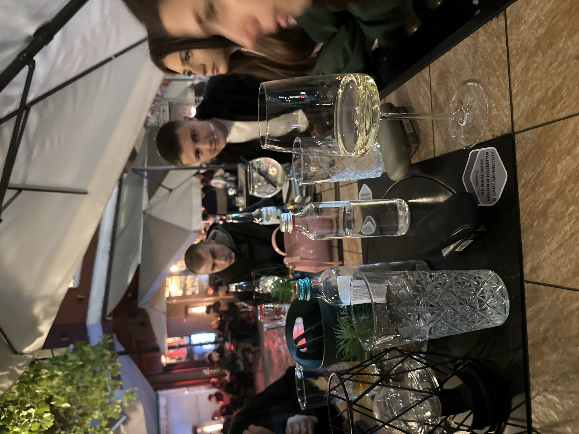The image shows a table setting at a restaurant in Plovdiv, Bulgaria. Three individuals are partially visible: two young women and a young man are seen near the top. The table holds a wine glass filled with a light-colored liquid, another patterned glass, two small water bottles, and a decorative metal container with greenery inside. There's also a small sign with Cyrillic text. Beyond the table, the outdoor seating area is shaded by white umbrellas, and the blurred background shows other diners and lights. The time of day is likely evening, given the lighting conditions. The pavement appears to be tiled in a light brown color.
