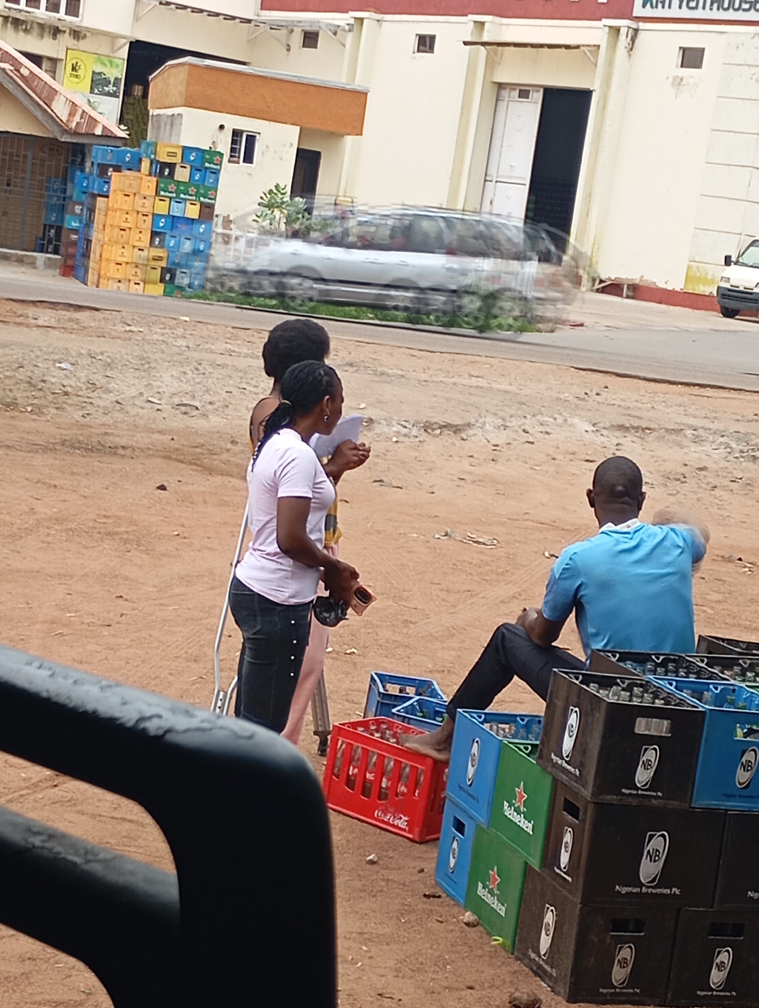 The image shows an outdoor scene in Shendam, Nigeria, with a group of people and stacks of crates. A building with a red roof and cream-colored walls is visible in the background, along with parked vehicles, some of which appear to be moving due to motion blur. In the foreground, three individuals are present. Two women are standing, with one holding a piece of paper and the other looking down. A man is seated, facing away from the camera. Several crates are stacked on the ground, some containing bottles. The ground is sandy and there is some debris scattered around. The lighting suggests it is daytime.