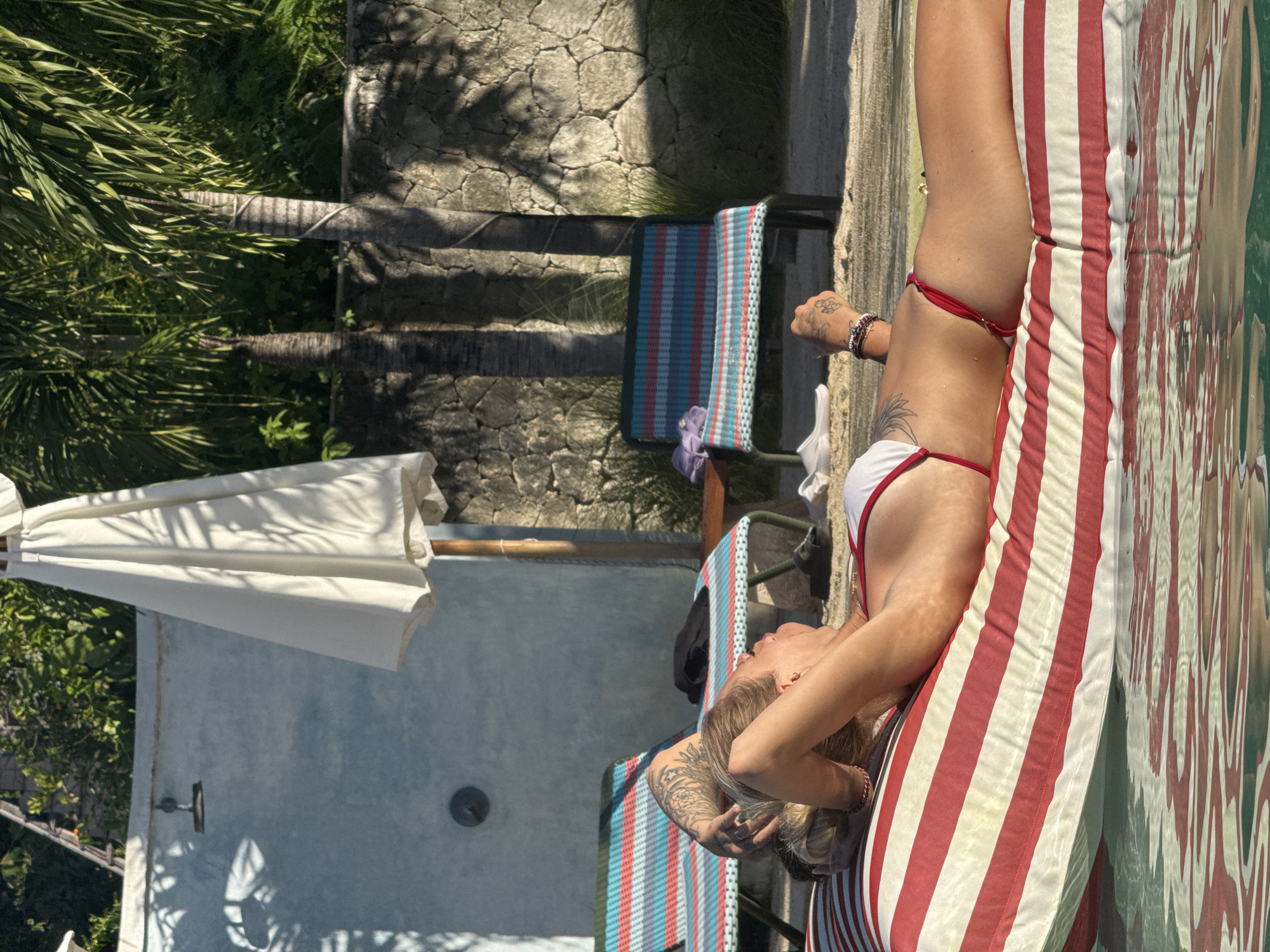 A fair-skinned woman with visible tattoos is reclining on a red and white striped beach chair near a swimming pool. The woman is wearing a white bikini with red trim. Her right arm is raised behind her head, her left arm is at her side. In the background, there are two beach chairs with blue and red stripes, an umbrella, and a stone wall with trees in the distance. The photo was taken during the day. The location is likely Nusa Penida, Indonesia, but this cannot be determined from the image alone.