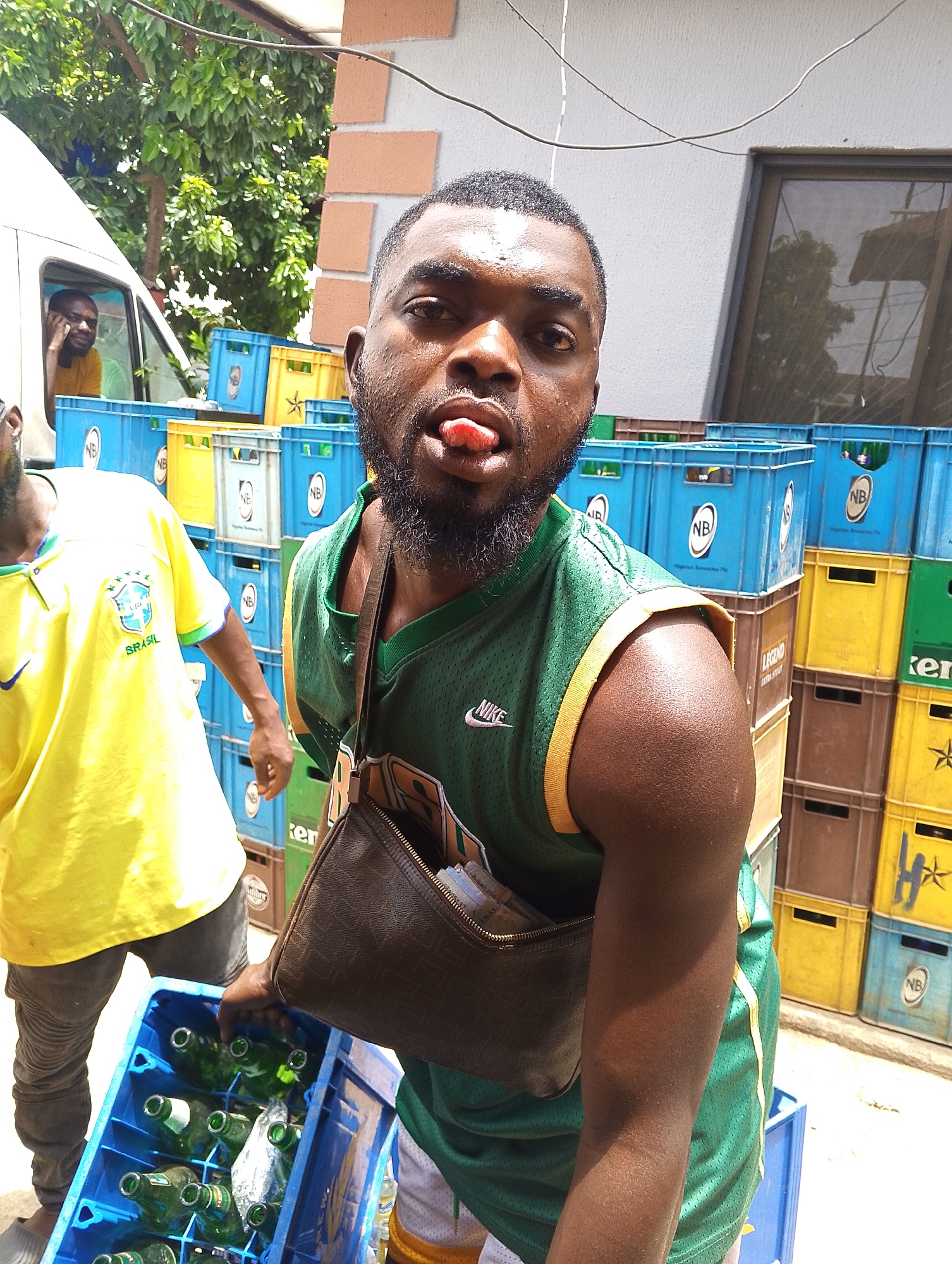 A dark-skinned man with a beard and short dark hair is prominently featured in the foreground, looking directly at the camera with his tongue extended. He appears sweaty, with visible moisture on his forehead and face. He wears a green and gold sleeveless mesh sports jersey with "NIKE" visible on the left shoulder. A brown leather-like cross-body bag or purse is slung over his shoulder. His right hand rests on the edge of a blue plastic crate filled with empty green glass bottles.
To the left, another dark-skinned man is partially visible, wearing a bright yellow t-shirt with "BRASIL" and a national team emblem printed on it. Further back, inside a white van, a third dark-skinned man is seen looking out the driver's side window.
The scene is outdoors under bright daylight, indicated by strong illumination and shadows. In the background, numerous plastic crates are stacked high. These crates are predominantly blue and yellow, with some brown and green ones. Many blue crates feature the logo "NB," while some yellow crates have a star emblem and the text "Heineken" or "LEGEND" is visible on other crates. Behind the stacks of crates, a white building wall with some reddish-brown brick accents is visible, along with green foliage from trees. Overhead, utility wires or cables crisscross. The overall setting appears to be an area for handling and storing beverage crates.