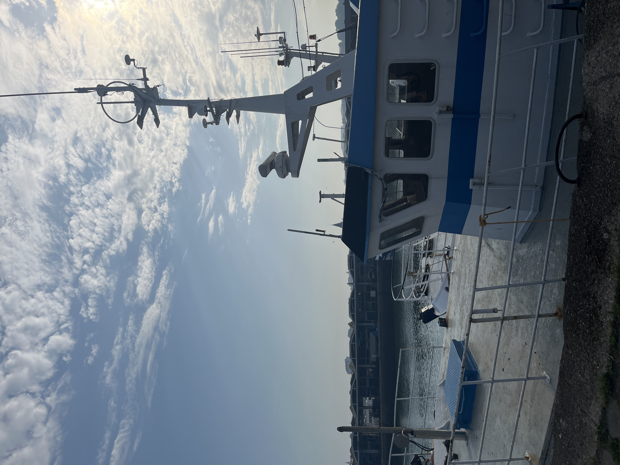 This is an outdoor, medium shot photograph of the deck of a white and blue fishing boat docked in the harbor of Dieppe, France. The boat is in the foreground on the right side of the image. Its deck is made of concrete and is surrounded by a white metal railing. A blue metal hatch is visible on the deck. The boat has a cabin with multiple windows. Above the cabin is a tall white mast with various antennas and equipment. In the background, across the water, is a row of buildings that appear to be warehouses or industrial structures. The sky is blue with scattered white clouds, and the sun is shining through them, creating a bright light. The water in the harbor is calm. There are no people visible in the image.