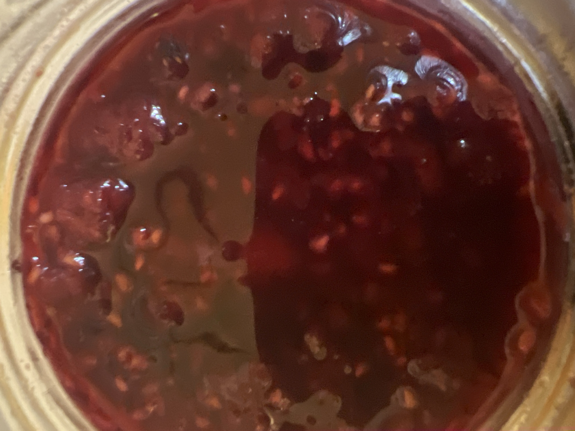 A close-up, overhead view of the surface of raspberry jam in a glass jar. The jam is a deep red color and appears to have chunks of raspberries visible throughout. The surface is slightly rippled and reflects light in a few areas, creating bright highlights. The edge of the glass jar is visible in the upper portion of the image.