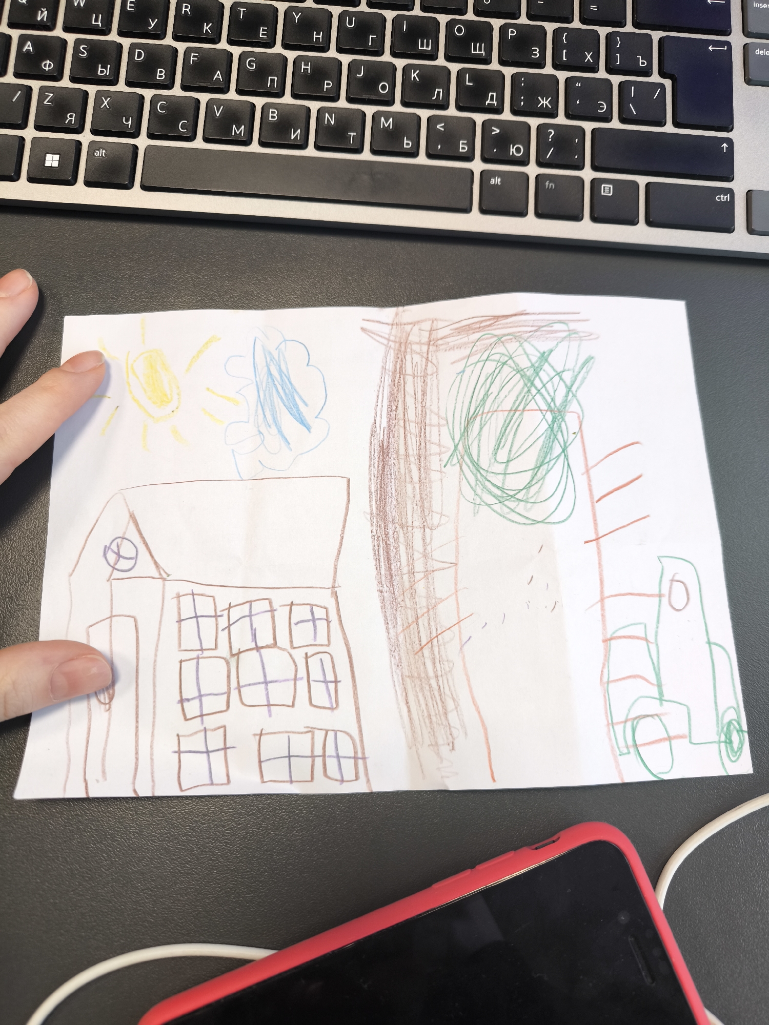 The image shows a child's drawing on a white piece of paper, placed on a dark grey surface with a laptop keyboard visible at the top. A portion of a hand with a finger pointing at the drawing is seen on the left side. A mobile phone in a red case with a white charging cable is at the bottom. The drawing depicts a simple scene with a brown house featuring a round shape above the entrance and multiple window shapes. Above the house, there are yellow sun rays and blue clouds. To the right, there is a brown drawing of a tree with green scribbles representing the leaves and grass. Further right, there appears to be a green vehicle.