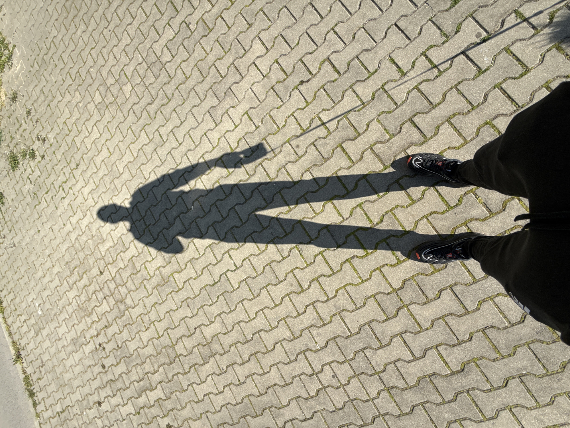The image shows a person's shadow cast on a paved surface. The person is wearing dark clothing and dark shoes with red and white accents. The paving stones are light gray and have a textured surface with some grass growing in the cracks. The shadow suggests the person is holding their right arm out to the side. The location is a paved area, possibly a sidewalk or pedestrian zone. There is no information to indicate this is located in Stara Zagora, Bulgaria.
