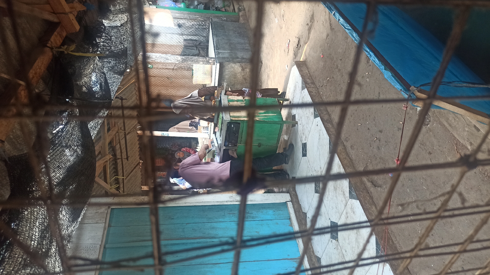 A man in a pink shirt and dark pants stands behind a green metal food cart, appearing to be preparing food or making a transaction. He is facing a woman wearing a hijab and a brown dress, who is standing on the other side of the cart. Behind them, a glimpse of a market-like setting is visible, with other people and structures. The scene is framed by a metal grate in the foreground. The location is identified as Trisinar, Indonesia.