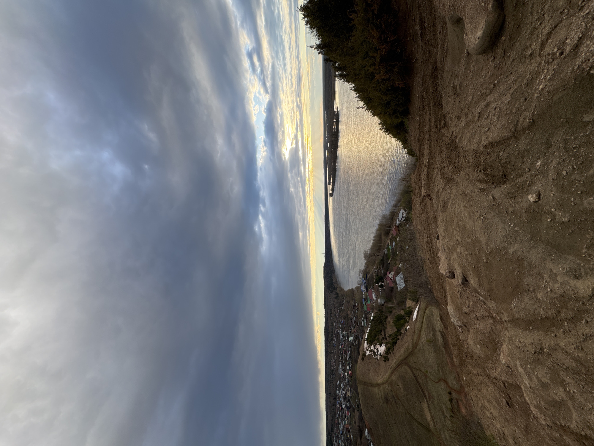 This panoramic image, correctly oriented, depicts a vast landscape from a high vantage point, looking down upon a river, a village, and a dramatic sky.
In the bottom-right foreground, a steep, earthy bank or cliff shows loose soil, scattered rocks, and sparse, dormant vegetation. Faint trails or paths are visible winding across the slope.
The midground is dominated by a wide body of water, likely a river, reflecting the low sun's golden light. Along the far bank, a village or small town is nestled, comprising numerous houses with various roof colors. Patches of snow on the ground indicate an early spring or late winter season, with trees still mostly bare. A forested area or more dense vegetation runs along the river's edge on the opposite bank.
The background features a expansive sky filled with diverse clouds. Darker, heavier clouds are prominent in the upper-left, while towards the horizon, brighter clouds reveal shafts of sunlight breaking through, creating a striking contrast and illuminating the water below. The time of day appears to be late afternoon or early evening, judging by the low, warm light.
No people or specific activities are visible. The scene conveys a sense of tranquility and the grandeur of nature, with the interplay of light, water, and clouds. There is no visible text. The location is Ольховка, Russia.