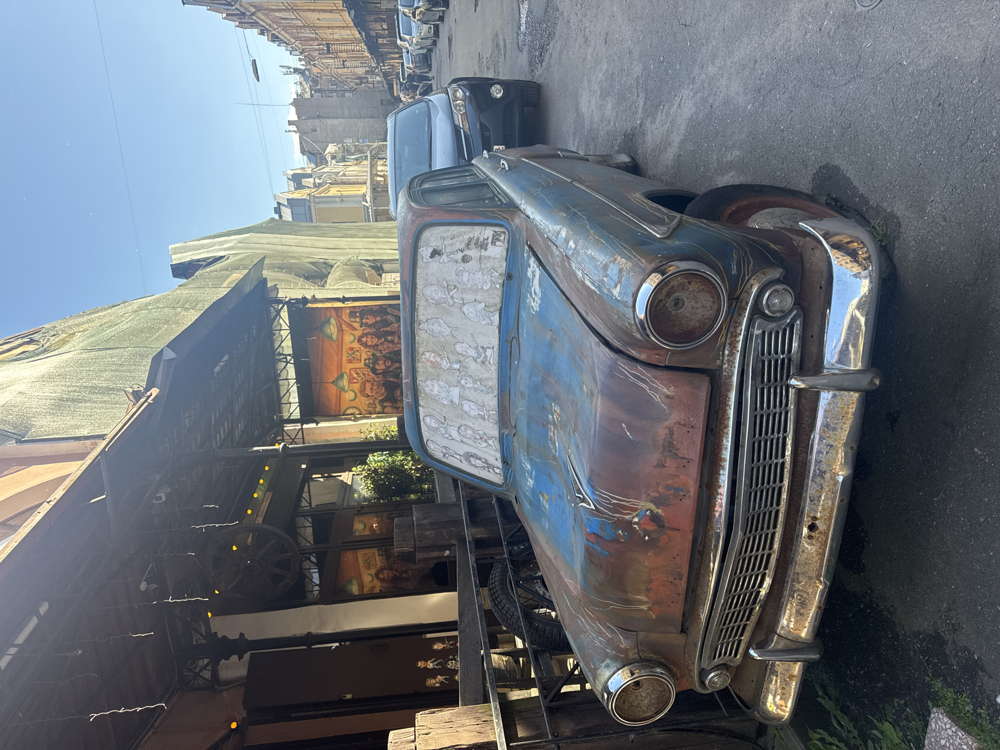 This image depicts a street scene in Saint Petersburg, Russia, under a clear, sunny sky, suggesting it is daytime. The primary focus is a weathered, vintage car, likely a GAZ-21 Volga, parked on an asphalt street. The car is a prominent subject, showing signs of rust and peeling paint, with a faded blue and red finish. Its chrome grille and bumpers are still somewhat shiny, hinting at its past glory. The windshield is covered with a decorative fabric featuring what appears to be a line drawing of cartoonish figures.
In the background, a line of other cars is visible, including a dark-colored SUV. The setting includes buildings with light-colored facades, some with ornate architectural details, characteristic of Saint Petersburg. To the left of the car, a building with a rustic facade is partly visible. It has wooden elements, possibly part of a cafe or shop, adorned with strings of yellow lights and a decorative cartwheel. Murals or artwork featuring stylized figures are visible on the walls of this building.
The overall mood is one of urban decay contrasted with hints of artistic expression and vibrant street life. The sunlight casts sharp shadows, reinforcing the sense of a bright, possibly warm, day. The scene captures a moment in the everyday life of the city, highlighting the juxtaposition of old and new, automotive history, and local urban aesthetics. There is no discernible text within the image.