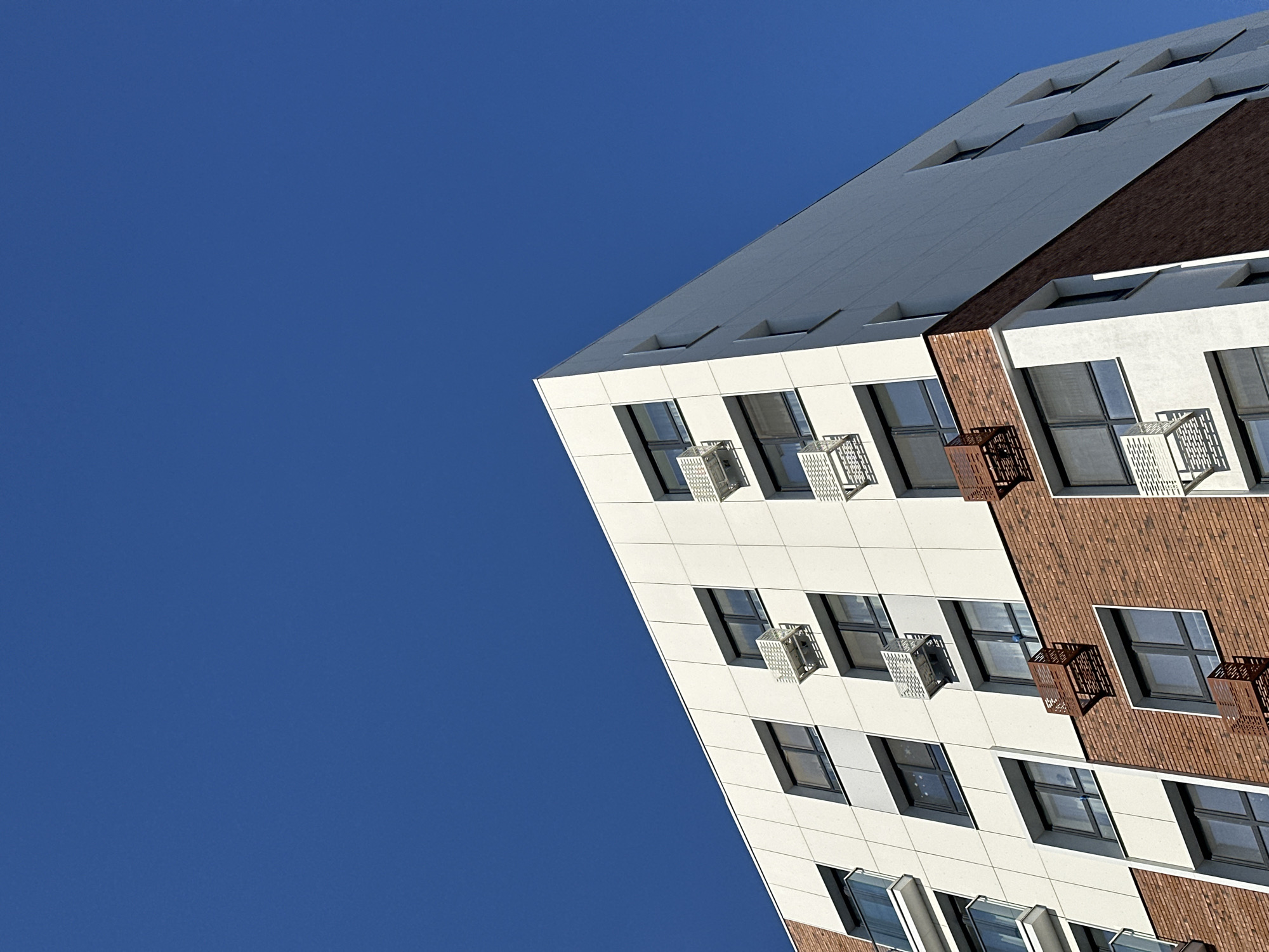 The image shows an upward view of a building facade in Kazan, Russia, against a clear blue sky. The building has a modern design with a combination of white paneling and brown brickwork. Windows are visible, some with small, ornate, open cages projecting outwards, possibly for air conditioning units or small balconies. The building appears to be multi-story, with a corner of the roofline visible. The composition is angled, giving a dynamic perspective.