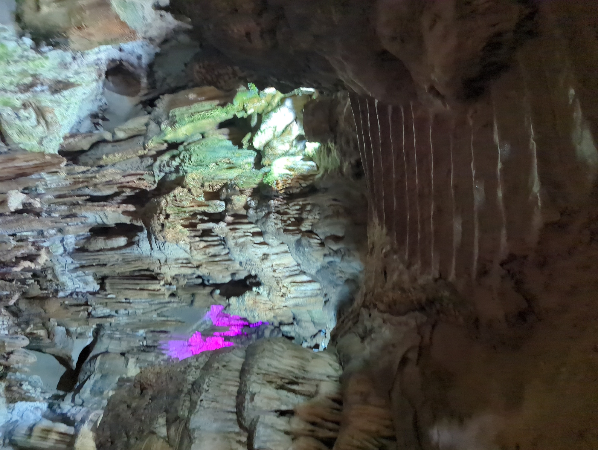 The image depicts the interior of a cave, likely a tourist attraction, with a pathway of stone steps leading upwards and into the cavern. The cave walls and ceiling are adorned with numerous stalactites and stalagmites, some illuminated by artificial lighting. Notably, a section of the cave is lit with vibrant purple light, creating a dramatic contrast with the natural earthy tones and the green lighting in other areas. The environment is rugged and natural, with rough rock formations dominating the scene. The image is focused on the cave's geological features and the lighting used to enhance its visual appeal.