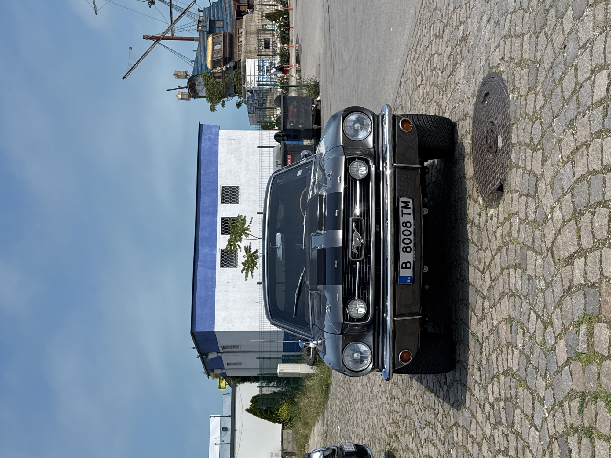 A vintage Ford Mustang, appearing to be a classic model from the mid-1960s, is prominently parked on a cobblestone street under a clear blue sky. The car is dark gray or silver with two distinct black racing stripes running down its hood and roof. It features chrome bumpers, a chrome grille with the Mustang emblem, and dual round headlights along with smaller, rectangular fog lights below the grille. The front license plate, visible on the bumper, reads "B 8008 TM" with "BG" indicating Bulgaria.
In the background, to the left of the car, stands a distinctive two-story building that resembles the hull and structure of an old wooden ship, complete with masts and rigging. Further to the left, a white building with a blue upper trim and visible windows is partly obscured. Palm trees are visible near this building. A few indistinct figures of people can be seen in the middle distance near the ship-like structure and some trash bins. The ground beneath the car and extending to the right is paved with rough, gray cobblestones, and a circular metal manhole cover is visible near the front right wheel of the car. The scene depicts a bright, sunny day, suggesting a casual outdoor setting, possibly a car show or a public parking area in a historical or port district.