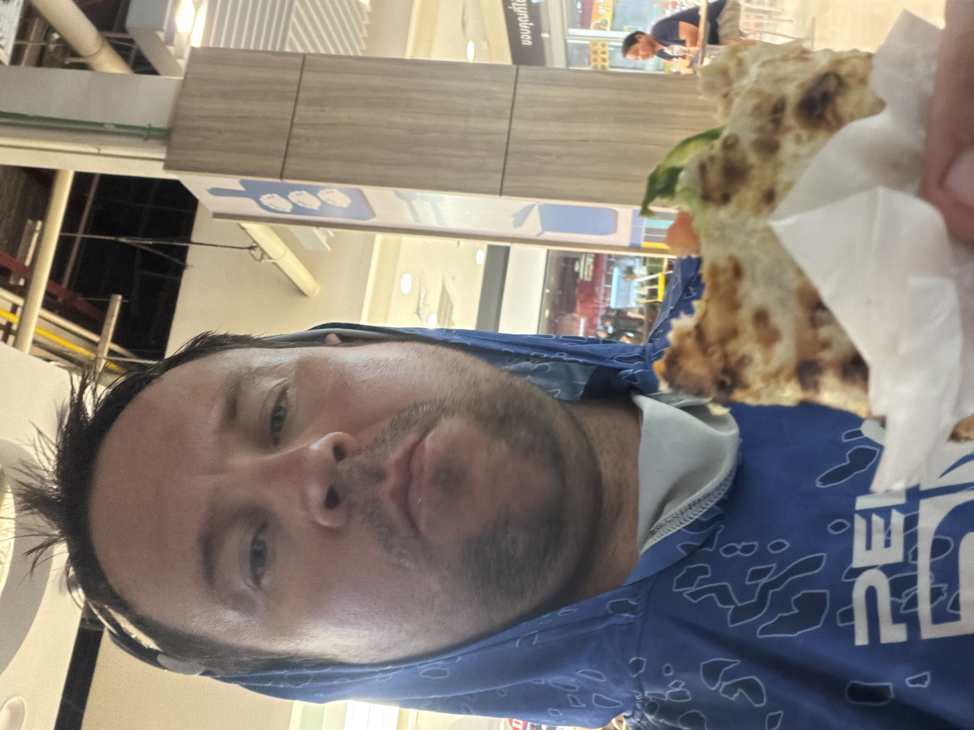 The image shows a man holding a partially eaten flatbread wrap up to his face for a selfie. He is wearing a blue hooded jacket with a white abstract pattern. His hair is dark and styled messily. He has a light beard and mustache. The background appears to be an indoor public space with fluorescent lighting, possibly a food court or shopping mall. There are pillars with signage, one of which is light brown and textured, and another with blue and white graphics of food items. In the background, to the right, a man in a dark blue shirt and shorts is seated at a table, looking away from the camera. The man in the foreground is the primary subject, and his expression suggests he is eating or about to eat the wrap. The image is taken from a low angle, common for selfies. The location appears to be in Ban Bang Rak, Thailand, as indicated by the sign in Thai script visible on a pillar in the background.