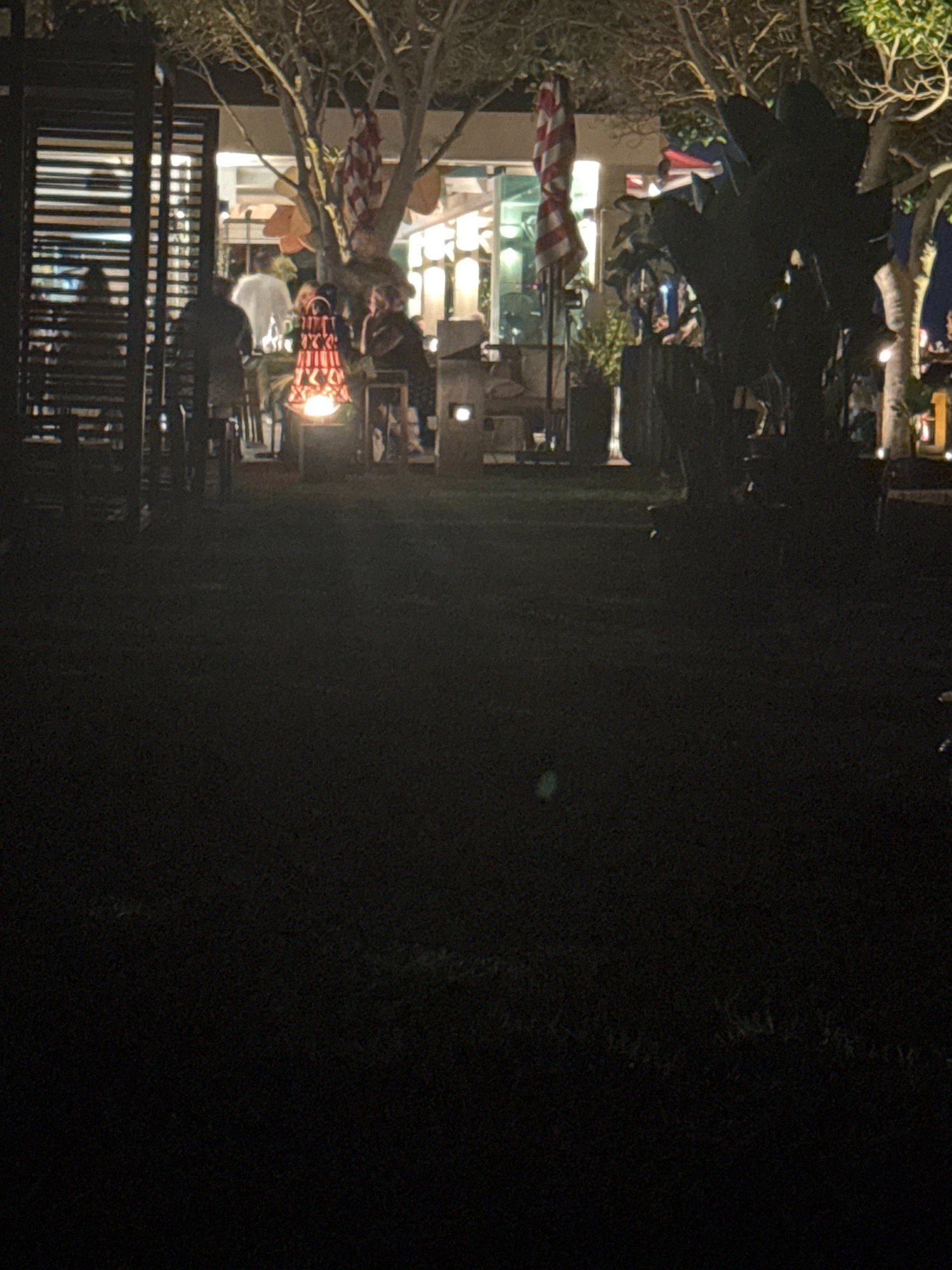 The image depicts an outdoor nighttime scene at a restaurant or bar in Agios Tychona Community, Cyprus. A group of people are gathered around a fire pit, which is emitting a warm glow and illuminating the surrounding area. The fire pit has a decorative, red metal surround. Two individuals are seated at a table to the right of the fire pit. In the background, there are tables and chairs, suggesting an outdoor seating area. Trees with sparse foliage are visible, and decorative lights are strung throughout the branches, adding to the ambiance. A striped umbrella is also visible, possibly indicating awnings or decor. The overall atmosphere suggests a relaxed evening gathering.