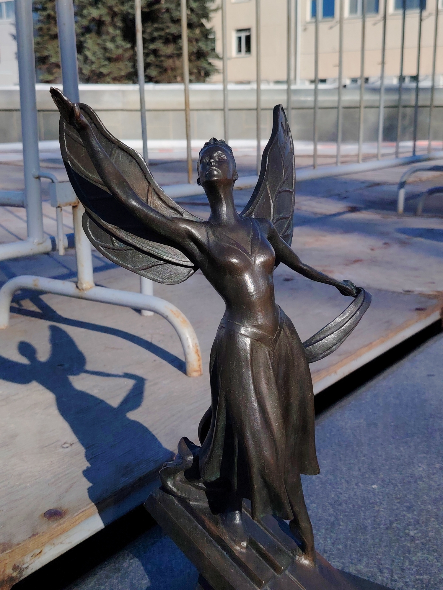 A full shot of a bronze statue of a winged woman located in the city of Chaikovsky, Russia. The statue depicts a woman with wings raised, holding a flowing fabric. She wears a dress and stands on a stylized base. The background includes a blurred urban setting with railings, trees, and a building with multiple windows. The lighting is bright, casting a distinct shadow of the statue on the surface below.