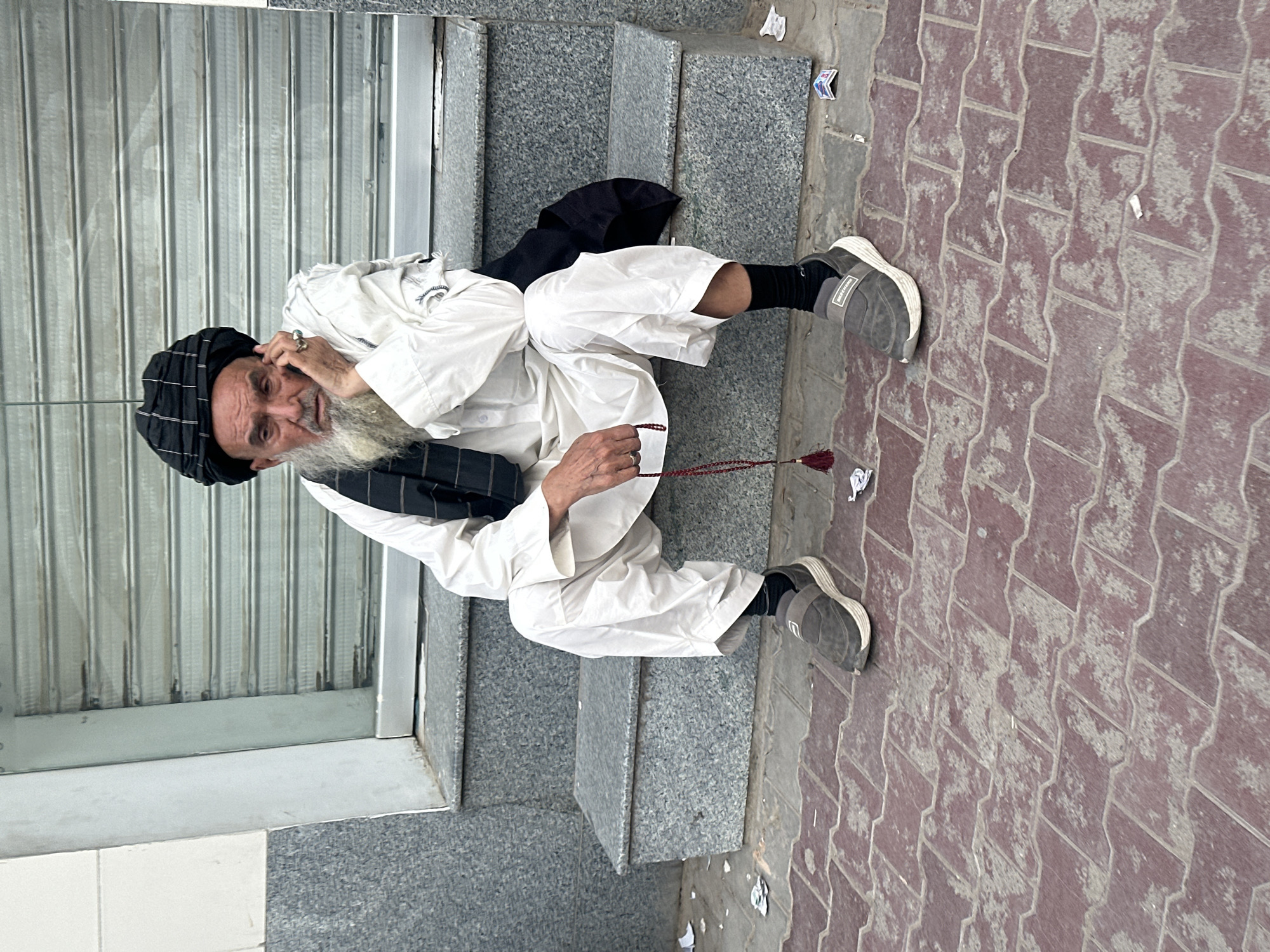 An elderly man with a long white beard and a black turban sits on a granite-clad step in front of a closed metal storefront. He wears a white tunic and trousers, with black socks and grey sneakers. A string of red prayer beads hangs from his right hand, and he rests his left hand on his face, looking towards the camera. Behind him, a grey metal roller shutter is visible. The ground in front of the step is paved with red brick-like tiles. There is some litter on the ground and on the steps. The location appears to be an urban street in Quetta City Tehsil, Pakistan.