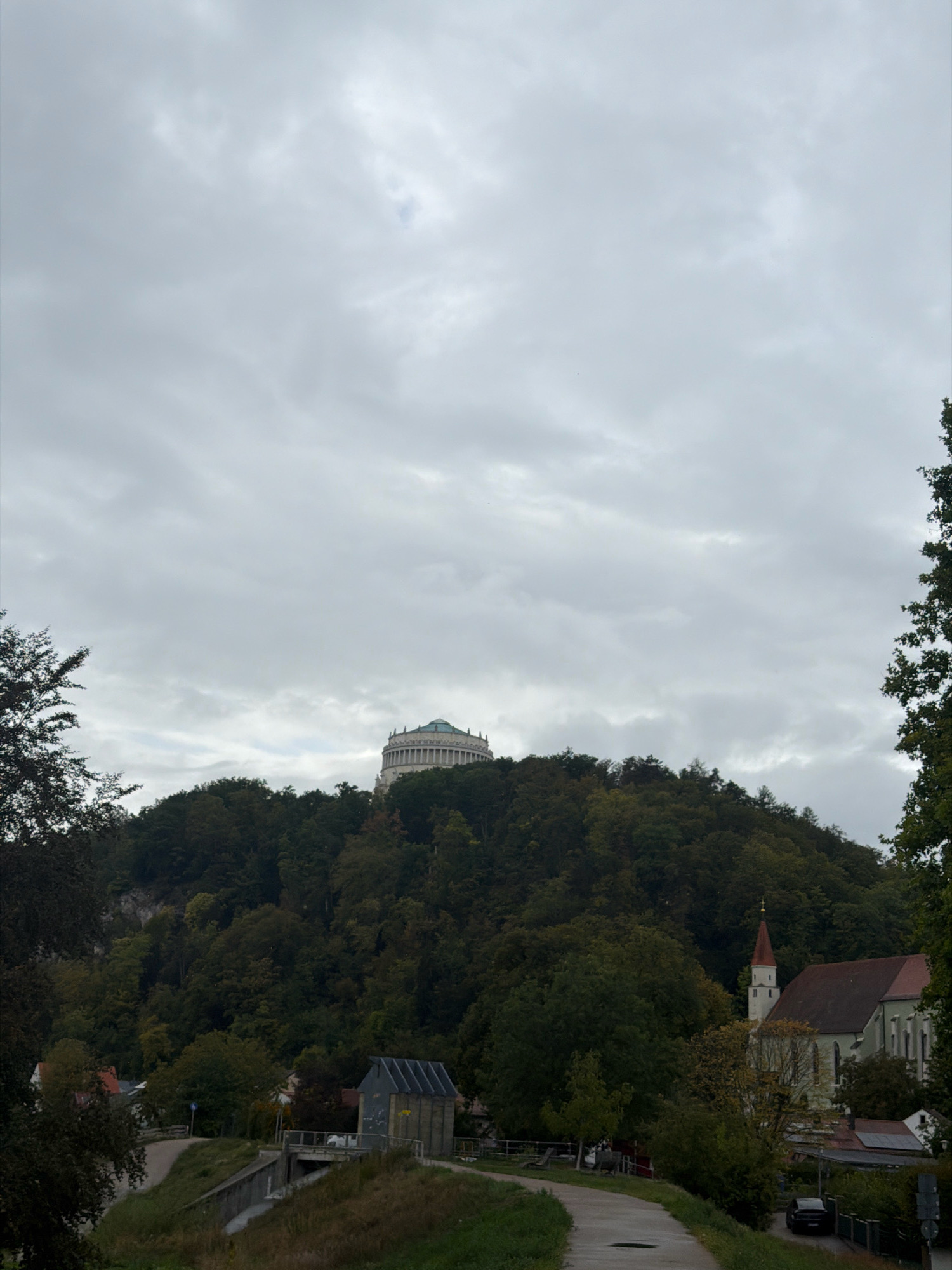 The image shows the Walhalla memorial building on a forested hill in the city of Kelheim, Germany, under a cloudy sky. In the foreground, a paved path winds through grassy areas. To the right, a church with a red-orange steeple stands among trees, adjacent to some residential buildings and a dark-colored car parked on the right side of the road. On the left, a small utility building with a metal roof is visible near a canal or bridge structure. The overall scene depicts a tranquil, overcast day in a scenic location with both natural and architectural elements.