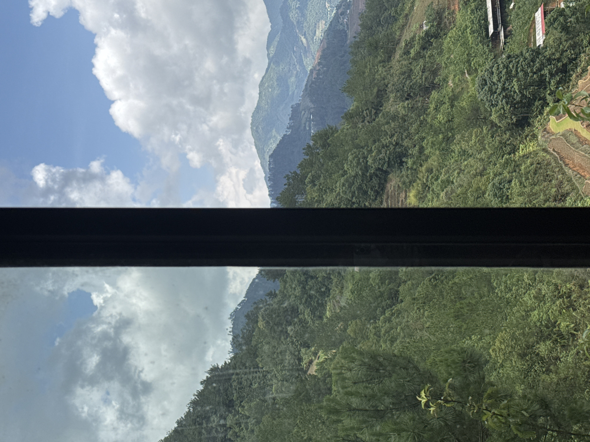 The image shows a view of a mountain range in Ngã Ba Kim, Vietnam, as seen through a window. The top half of the image captures the mountainous landscape and a cloudy sky. The bottom half is a mirrored reflection of the same scene on the window surface. The horizon line is bisected by a dark, rectangular frame, most likely a window frame. The mountains are covered in lush green vegetation, and the sky is a mix of blue and white, with thick clouds.