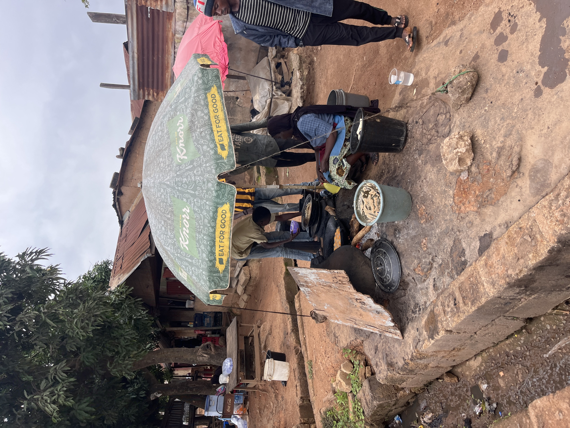 In Bukuru, Nigeria, an outdoor food stall is depicted under a large green Knorr umbrella and a smaller pink umbrella. Several individuals are present. A woman in a blue patterned dress is seated near the cooking area, which features a large, raised brick structure with cooking pots and a fire. Men stand around the cooking area, with one appearing to be preparing food and another observing. In the background, there are rudimentary structures with corrugated metal roofs, and trees. Various containers, buckets, and cooking implements are scattered around the stall. To the left of the cooking area, a small, muddy ditch is visible, containing debris. A wooden table with a cooler and some containers is situated further in the background.