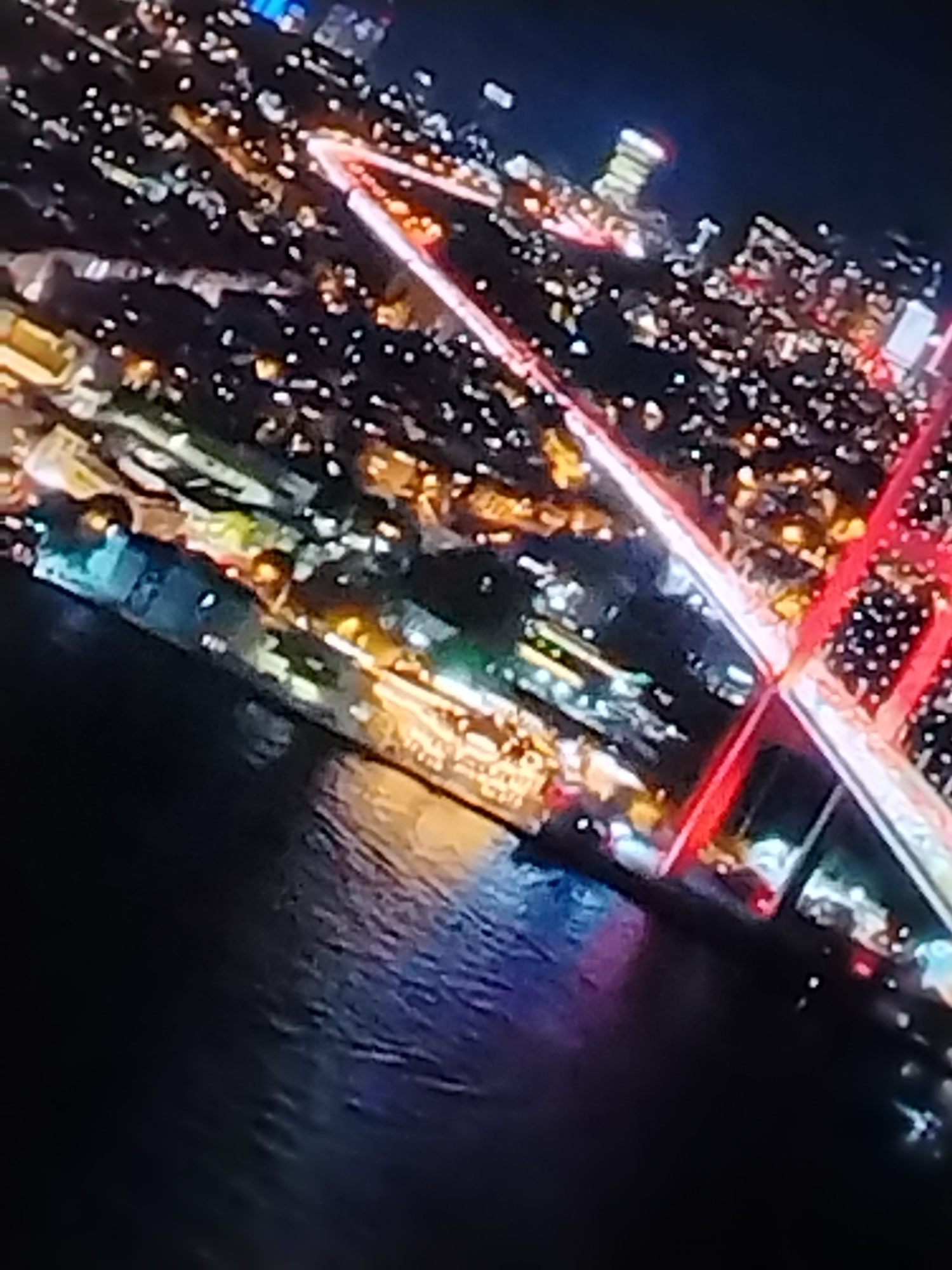 The image shows an aerial view of a city at night, with numerous lights illuminating the urban landscape. A body of water reflects some of the light, creating a shimmering effect. A bridge with distinctive red illumination spans a part of the water. The composition suggests a high vantage point, likely from a tall building or aircraft. The specific location is unknown.