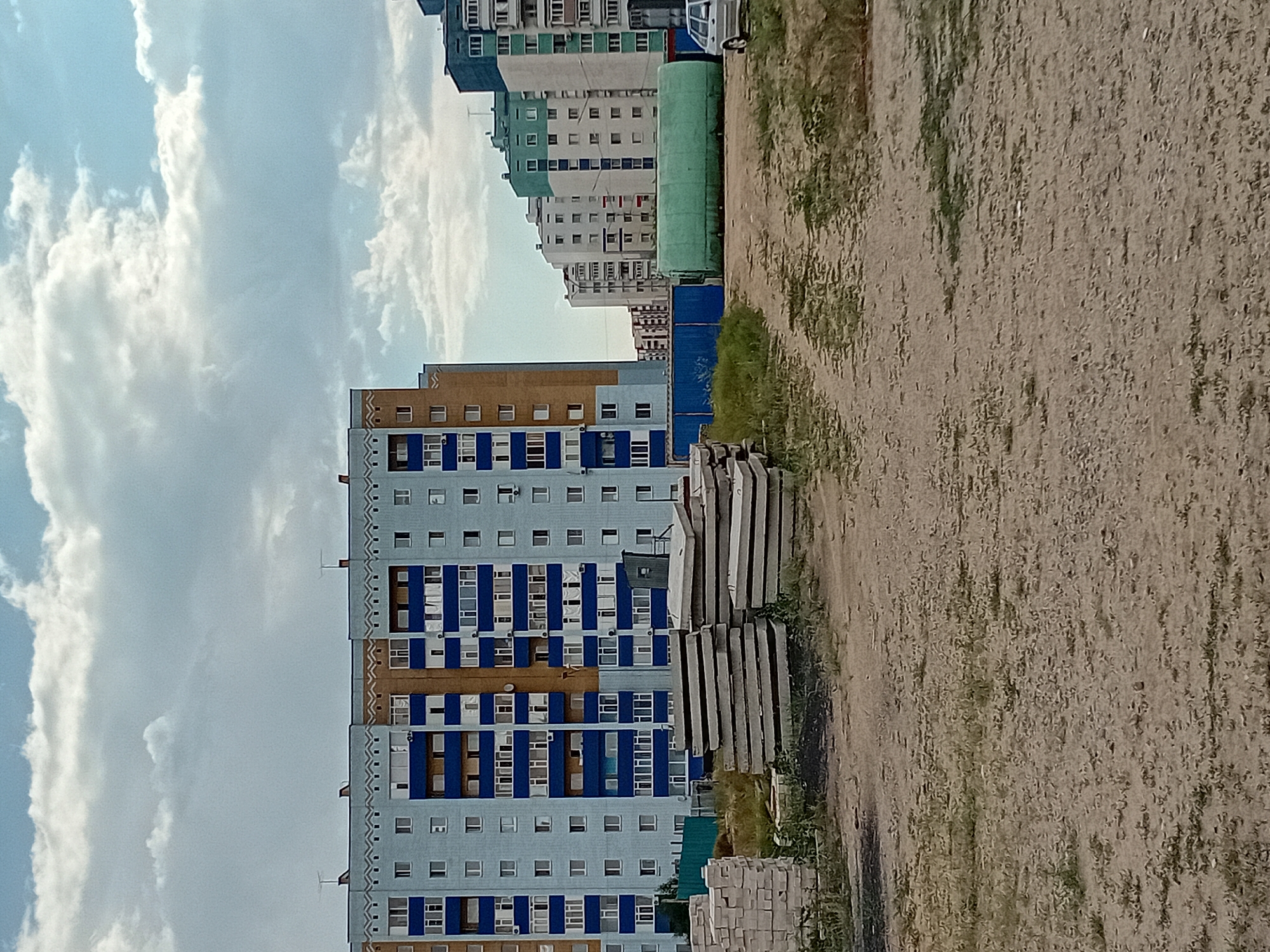 This is an exterior, eye-level, medium shot of a construction site in the city of Semey, Kazakhstan. The scene is dominated by several multi-story apartment buildings in the background, featuring light blue and orange facades with dark blue window frames. In the foreground, stacks of concrete slabs are piled on a dirt and grass area. A blue corrugated metal fence runs along the middle ground, behind which is a large, cylindrical green tank. A silver sedan is parked to the right of the green tank, and a pile of light-colored bricks is visible on the far left. The sky is partly cloudy with patches of blue. There are no people visible in the image. The overall impression is one of ongoing construction or development.