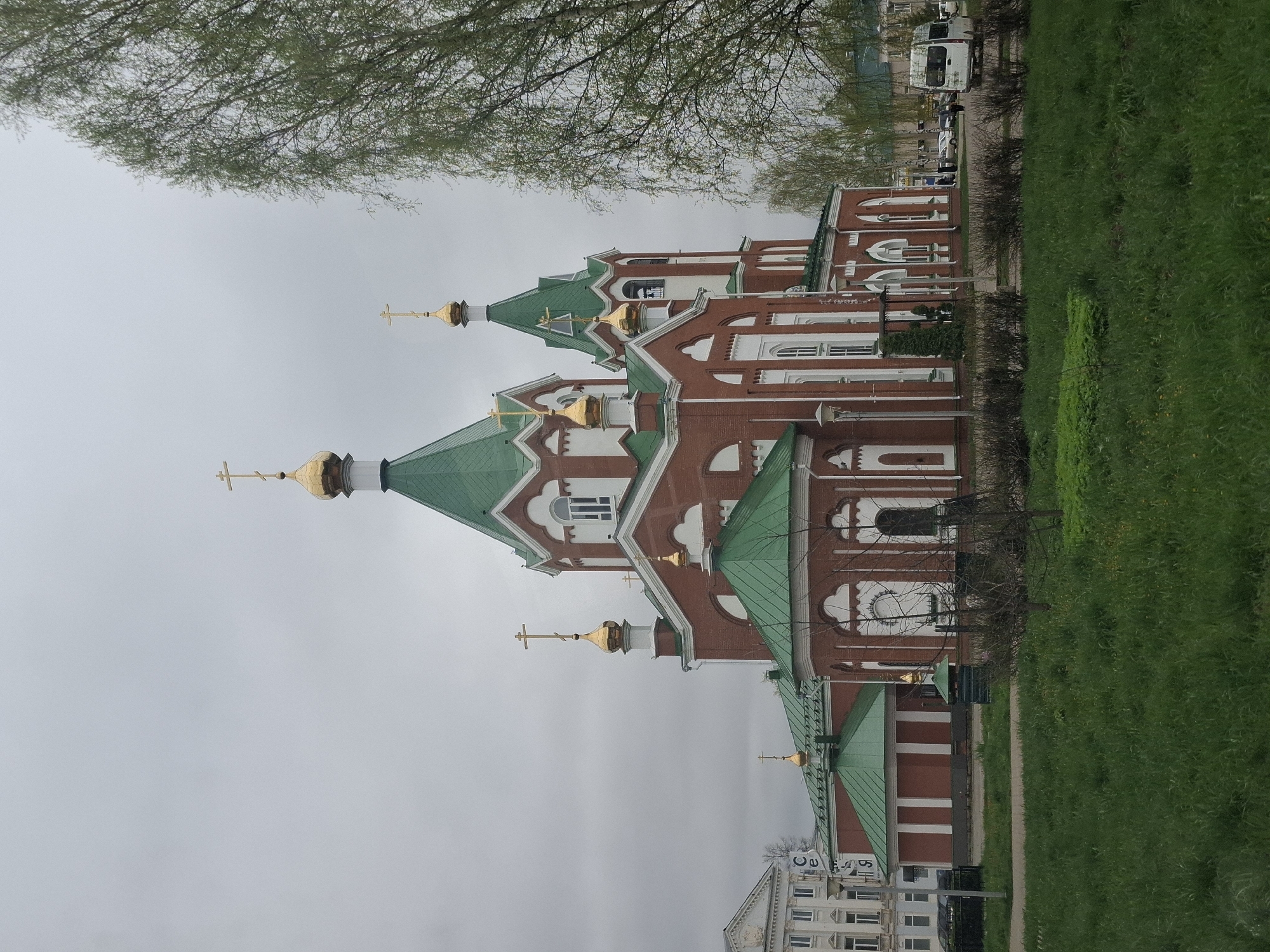 The image depicts the Alexander Nevsky Cathedral in Glazov, Russia. The cathedral, constructed of red brick with white architectural detailing and green roofs, has multiple golden domes topped with crosses. A patch of green grass lines the right side of the frame. A tree is visible in the upper-left corner. The sky is overcast. A multi-story building is visible in the lower-left corner and cars are on the street behind the cathedral.