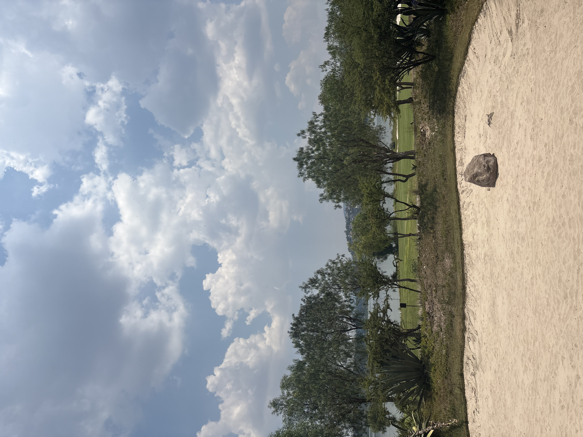 The scene is a sand trap on a golf course in Pueblo Nuevo de Jasso, Mexico. The trap is filled with light-colored sand, and a dark rock is visible within the sand. Beyond the trap, there is a strip of green grass, trees, and a body of water. The sky is cloudy. No people are visible.