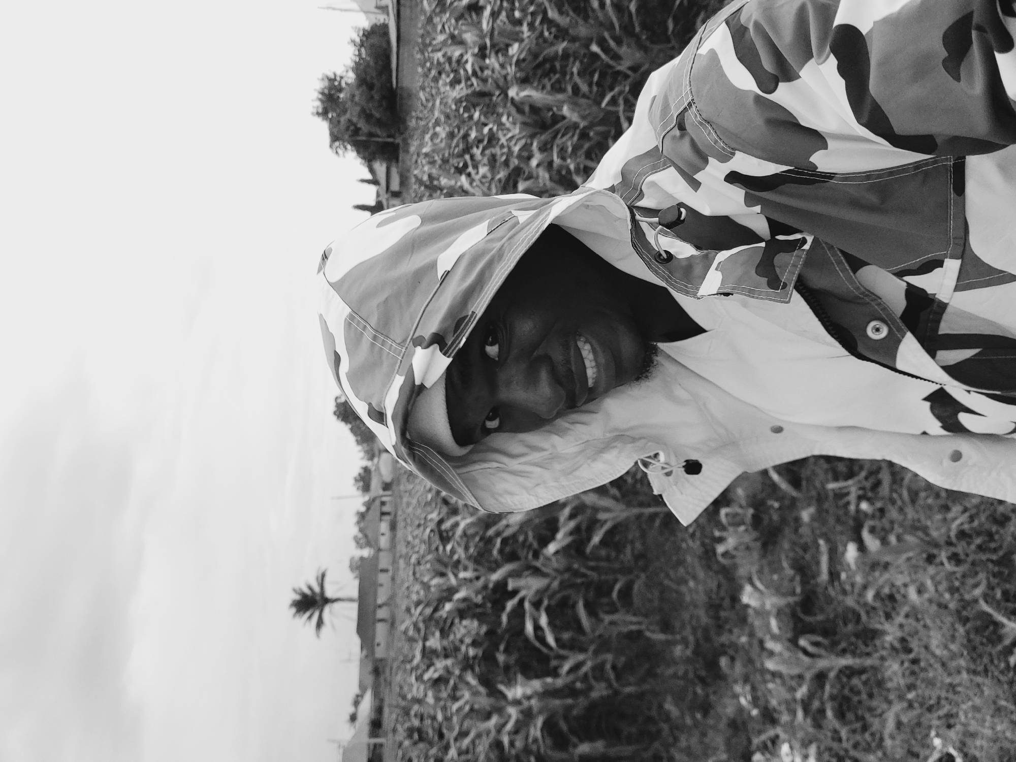 A young person with dark skin is captured in a close-up, outdoor portrait, smiling brightly towards the camera. He is wearing a hooded jacket featuring a white, black, and grey camouflage pattern, with the hood pulled up and framing his face. His left eye is visible, and his mouth is open in a wide, cheerful smile, revealing his teeth. He has visible facial hair, appearing as stubble or a short beard.
The background suggests an open, possibly rural or semi-rural, environment. Behind the person, a dense field of tall, leafy plants, resembling crops such as corn, stretches across the midground. Further in the distance on the left, a distinct palm tree silhouette stands against a bright, slightly hazy sky, which fills the upper and left portions of the frame. Faint outlines of other trees or structures are visible on the horizon. The bright illumination indicates it is daytime. The subject's expression conveys happiness or amusement. No text is visible in the image. The scene aligns with a location such as Bukuru, Nigeria.