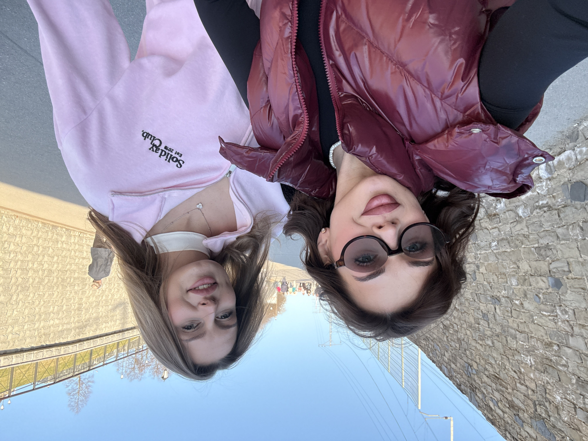 Two women are posing for a selfie. The woman on the left is wearing a pink zip-up hoodie. The woman on the right is wearing sunglasses and a puffy burgundy jacket. In the background, there is a group of people walking on a pedestrian bridge over the Kama River. The bridge is the Perm Bridge in Perm, Russia. The weather appears to be sunny.
