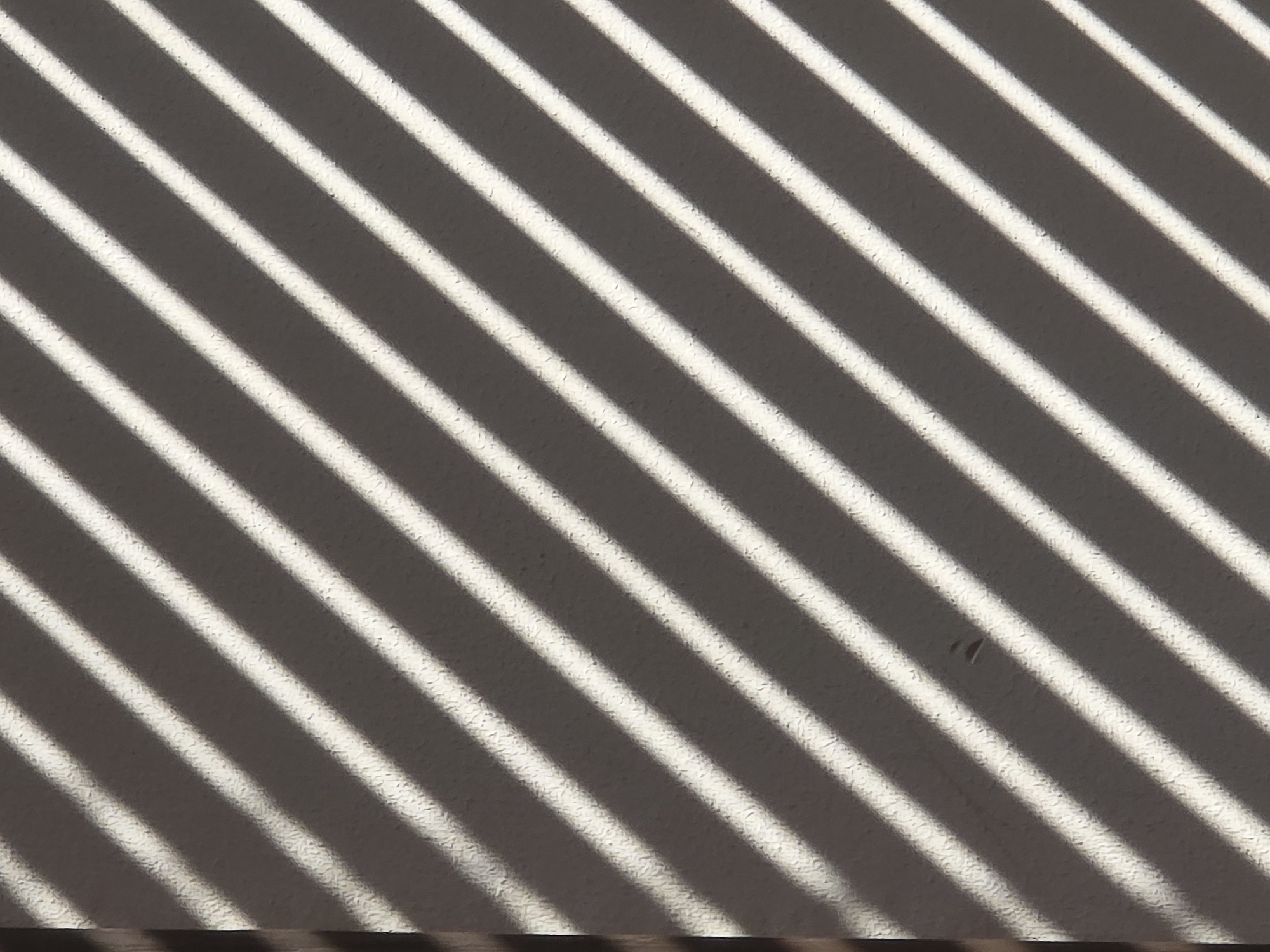 The image is a close-up, abstract shot of light and shadow patterns created by blinds on a textured, neutral-colored wall. Diagonal stripes of bright white light alternate with dark gray shadows, creating a linear, graphic effect. The wall texture appears somewhat rough, with visible stucco-like bumps. The lighting suggests it is daytime, casting distinct, sharp-edged shadows. There are no people or discernible objects within the frame, aside from a small, dark speck possibly a tiny insect or debris on one of the lighter stripes, and a vertical dark line on the far left edge of the frame which may be the side of the blind mechanism or a frame. The overall impression is one of a visually striking, minimalist composition formed by everyday elements. The image is devoid of any specific geographical or environmental context that would place it in a particular city or location.