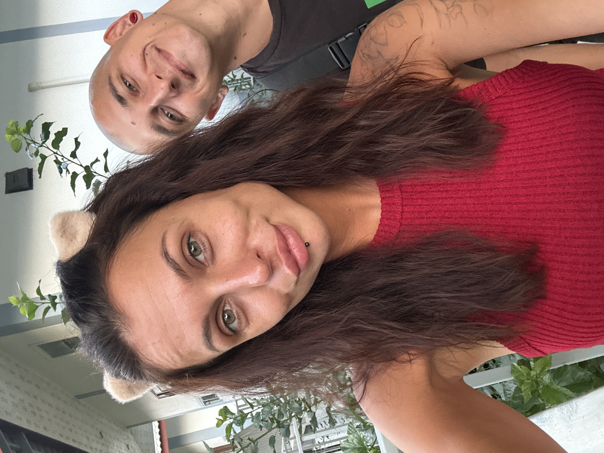 Two individuals, a woman with brown hair and tan cat ear headband, and a bald man with a red ear plug, are taking a selfie outdoors. The woman is in the foreground, wearing a red ribbed top, and has a lip ring. The man is partially visible behind her on the right, smiling. They appear to be on a balcony or outdoor seating area, with greenery and a white railing visible. A building with a tiled roof is in the background. The lighting suggests it is daytime. The location is Ko Samui District, Thailand.
