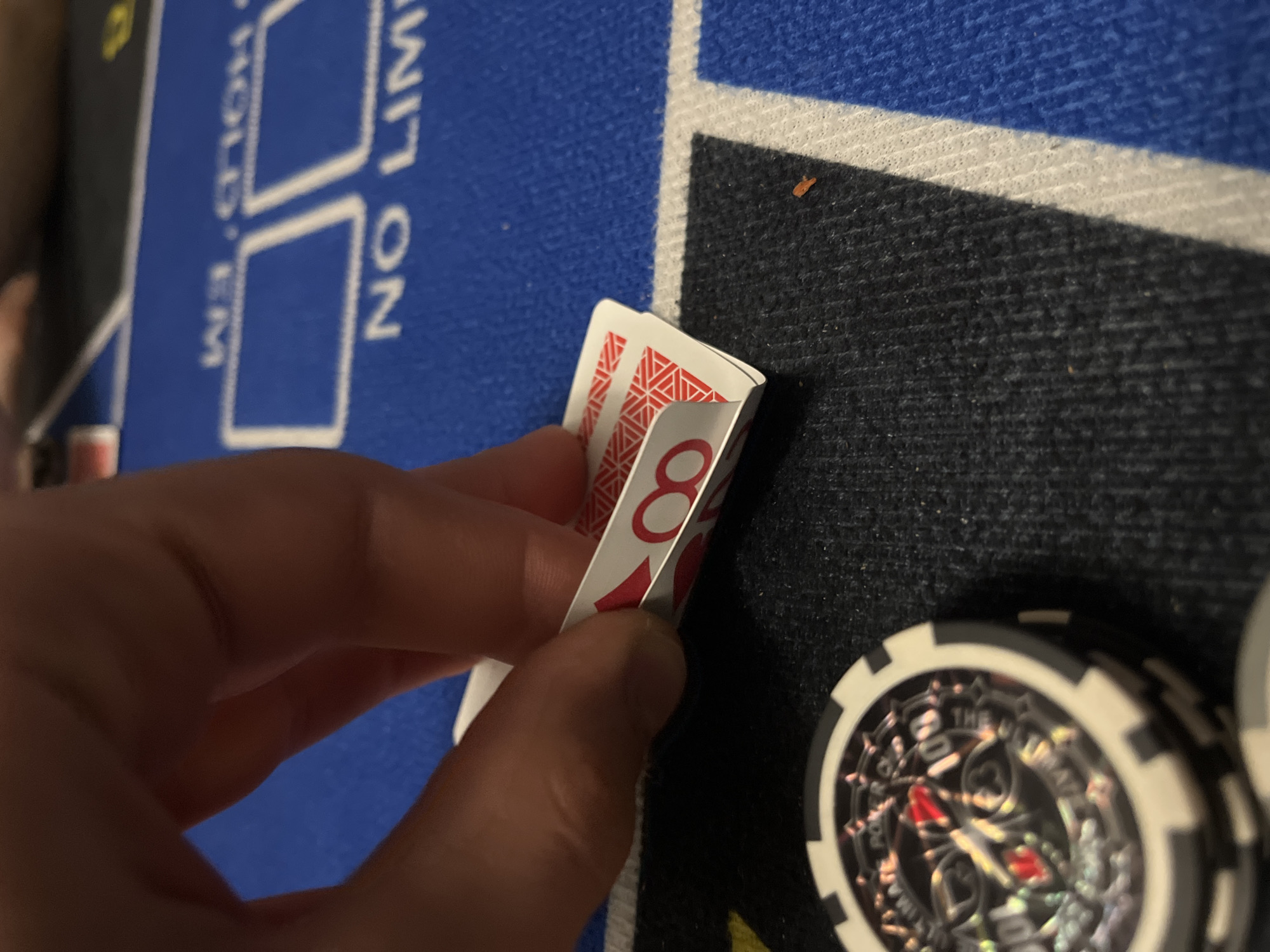 This close-up shot captures a moment during a poker game, showcasing a person's hand as they discreetly peek at their playing cards. The hand, with fingers curled around the edge of two cards, gently lifts them to reveal a corner of the top card. This card displays a red '8' along with a red suit symbol, likely a heart, against a white background. The reverse side of the cards features a repeating red and white geometric pattern.
The game is taking place on a poker table, indicated by the felt surface. A vibrant blue section of the table is visible on the left, featuring white text that clearly reads "NO LIMIT HOLD 'EM" and outlines of rectangular card placement areas. To the right, a darker, possibly black, section of the table felt is separated by a white stripe.
In the bottom right corner, a stack of poker chips is partially visible. The topmost chip is predominantly white and black, with a detailed, possibly holographic, metallic center. This intricate design includes various card suit symbols (hearts, diamonds, clubs, spades) and text, with "THE ULTIMATE" and the value "100" being discernible.
The scene suggests an indoor setting, possibly a casino or a home game, with artificial lighting, though specific time of day or location cues for Burgas, Bulgaria, are not present within the image itself. The focus is on the anticipation and strategy inherent in a poker game as a player evaluates their hand.