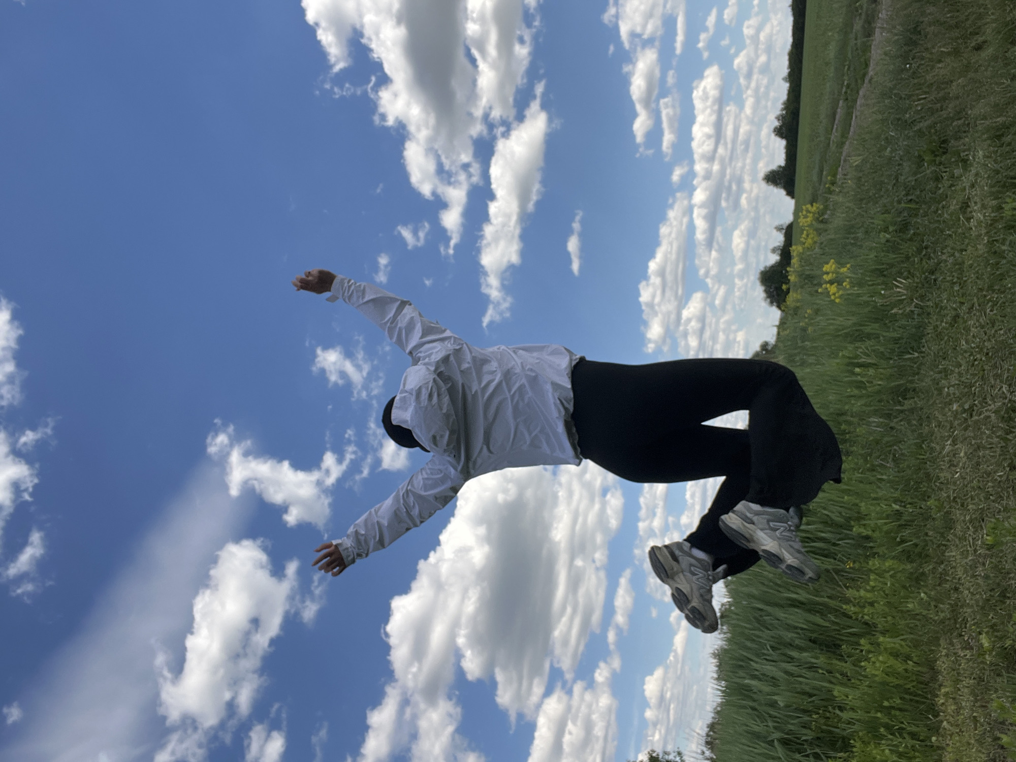 A person in black leggings, a white jacket, and gray sneakers is standing in a grassy field with arms outstretched. The sky is visible behind the person, filled with white, puffy clouds and patches of blue. The location appears to be outdoors, likely in a rural or natural setting. The specific location in Rudky, Ukraine, cannot be determined from the image alone.