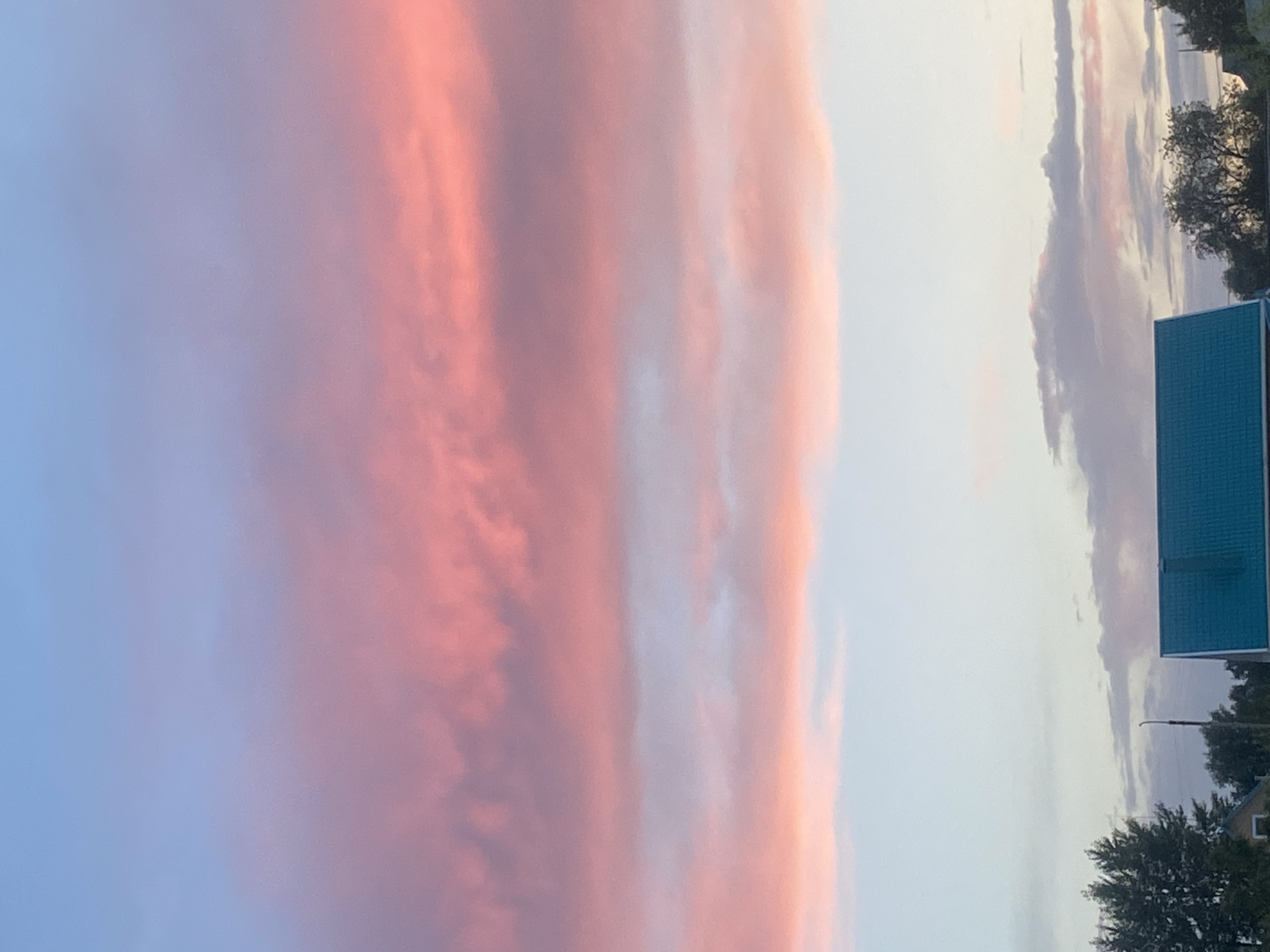 This is an eye-level, medium shot of a twilight sky over a rural residential area. The sky displays a gradient of soft blues and pinks, indicative of sunset or sunrise, with a prominent band of pink clouds stretching horizontally across the mid-frame. Below the sky, the upper portion of a teal-colored roof is visible in the lower right quadrant, belonging to a house. To the left of the roof, a streetlamp stands tall, and behind it, dark green trees frame the left edge of the image. The overall environment appears to be quiet and serene. The location is identified as Tolbazy, Russia. There are no people or animals visible in the scene, nor any discernible actions or interactions.