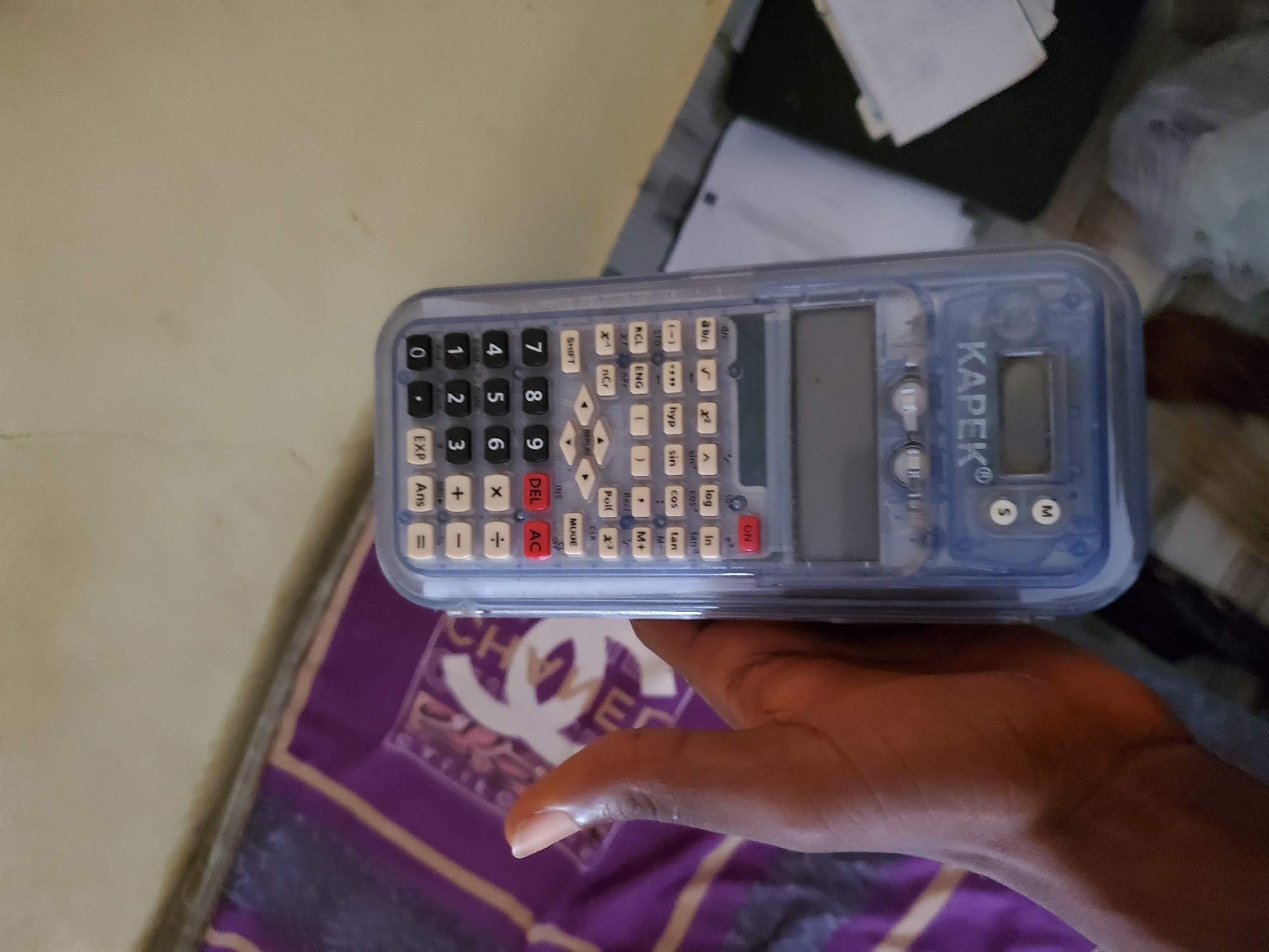 A person's left hand is holding a blue transparent scientific calculator in a close-up, eye-level shot. The calculator is positioned in the center of the frame, with its buttons and display visible. The background shows a patterned fabric, possibly a bedsheet or a rug, with a prominent white Chanel logo. The scene appears to be indoors. The image was taken in Jalingo, Nigeria.