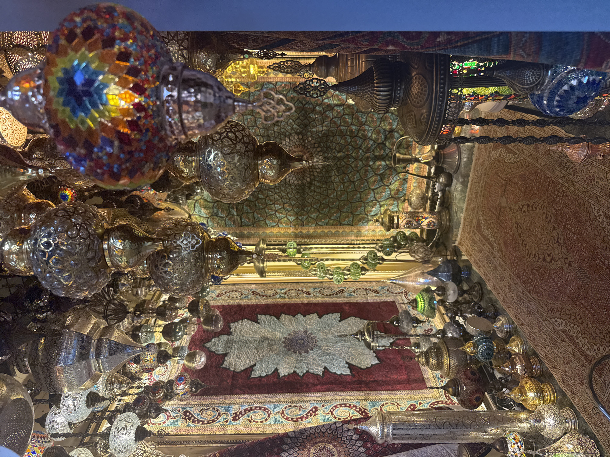 This detailed image captures the interior of a traditional Turkish lamp and carpet shop, likely situated within Istanbul's Grand Bazaar (Kapalıçarşı). The scene is densely packed with a vast assortment of ornate lamps and lanterns, predominantly made from hammered metal (brass and silver-toned) and multicolored mosaic glass, suspended from the ceiling at varying heights. A prominent mosaic lamp in the right foreground displays a vibrant, rainbow-colored geometric pattern. The shop's walls are draped with large, intricately patterned carpets or tapestries; one features a deep maroon field with a large, central, light-colored leaf-like motif, while another exhibits a green and blue pattern with a repeating circular design. The floor is covered by a traditional red and beige patterned rug. Along the lower left and right, various metal artifacts, including pitchers, bowls, and smaller decorative lamps, are arranged on counters or shelves. The illumination primarily originates from the numerous lit lamps, casting a warm and inviting glow throughout the space. No individuals are present in the scene.