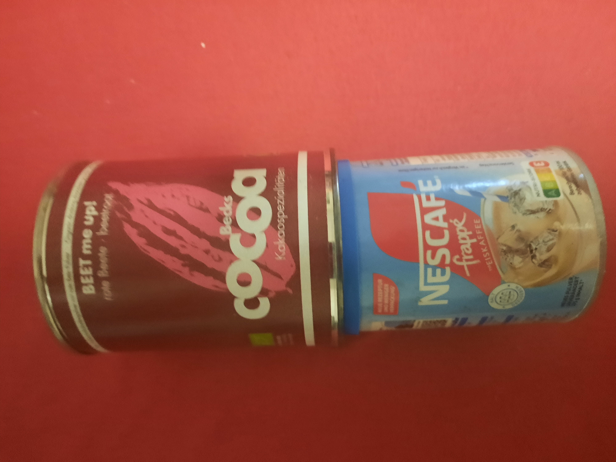 The image shows two cans of food products stacked on top of each other against a red background. The top can is maroon with white text and a pink graphic of a beet. It reads "BEET me up!" and "rote Beete - beetroot". Below this, it says "Becks Cocoa" and "Kakao-Spezialitäten". The bottom can is blue with white and red text and graphics. It reads "NESCAFÉ frappé" and "EISKAFFEE". There is also a graphic of a glass with a beverage and ice. The cans are positioned in the center of the frame. The background is a uniform red color. There are no people or other objects visible. The scene is indoors. The image does not contain any information about the city of Rosenheim, Germany.
