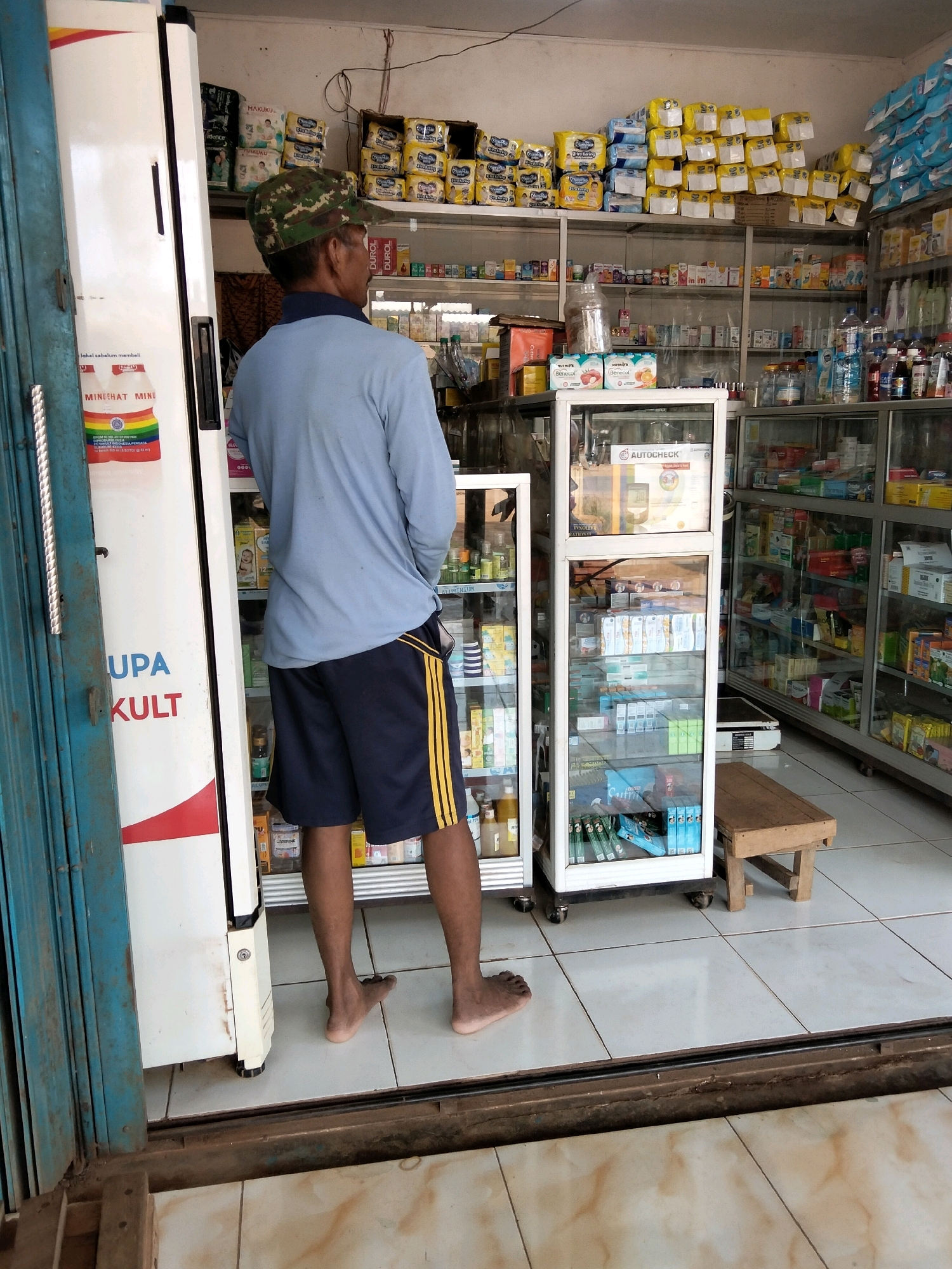 A man wearing a camouflage baseball cap, a light blue long-sleeved shirt, and navy blue shorts with yellow stripes stands with his back to the camera, facing away from a refrigerator and towards shelves stocked with various products in what appears to be a small convenience store or pharmacy in Sidomulyo, Indonesia. The man's feet are bare. To the left of the man is a white refrigerator with a red and white logo on its side. Behind the man, several shelves are filled with boxed items, including diapers, medications, and personal care products. In the foreground, a blue metal gate is partially visible. The floor is tiled with white and beige squares. A small wooden stool is positioned near a glass display cabinet filled with more products. The overall environment suggests a commercial establishment with a variety of goods for sale.