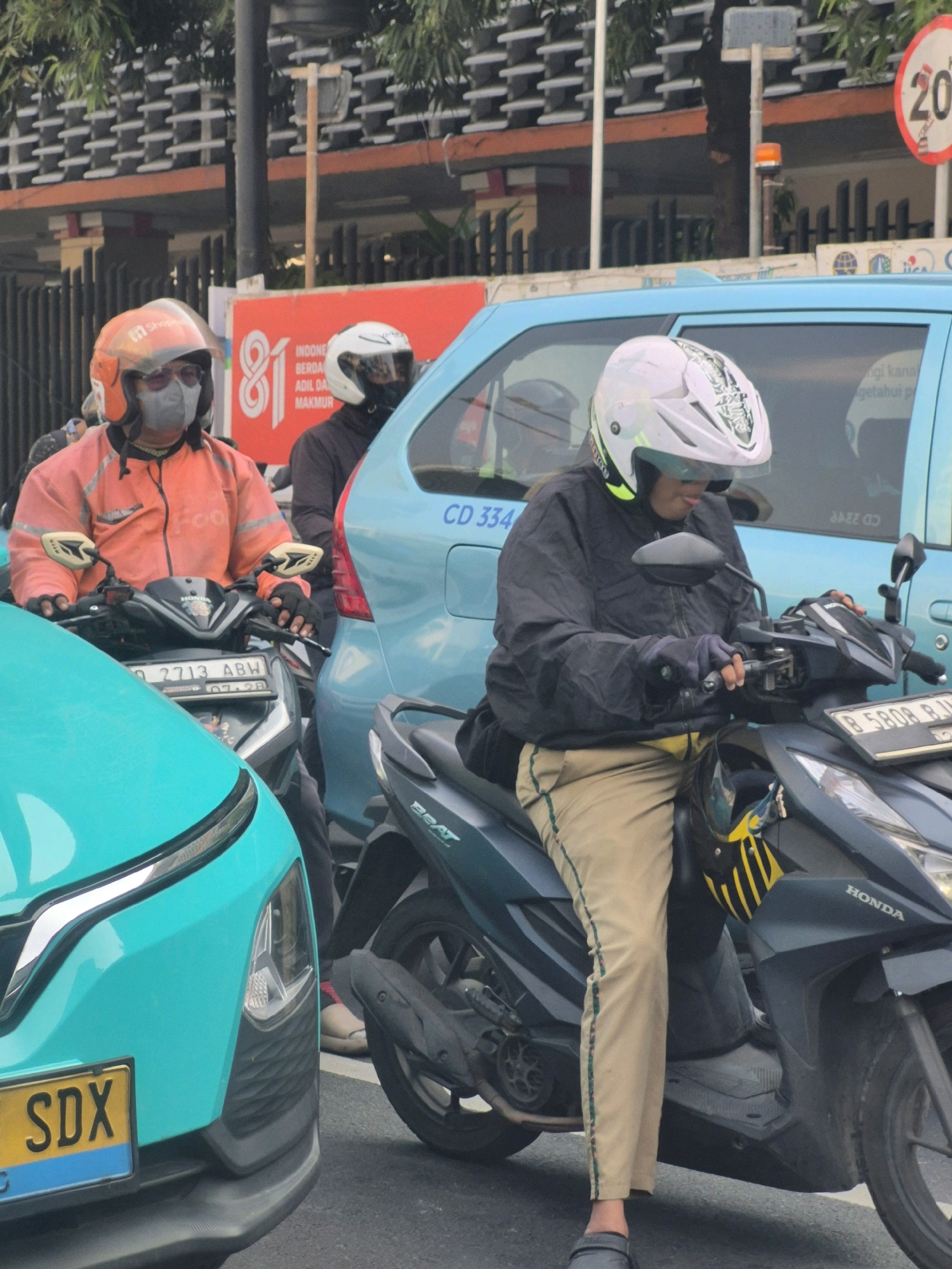 A medium shot captures a traffic scene in Central Jakarta, Indonesia, with several vehicles and people. In the foreground, a teal car is partially visible on the left, with a yellow and blue license plate reading "SDX". To its right, a dark gray motorcycle is ridden by a person wearing a white helmet with green accents, a dark jacket, and light brown pants with a green stripe down the side. They are holding the handlebars and looking down. Behind this motorcycle, another dark gray motorcycle is seen, with a rider wearing an orange jacket, grey mask, and orange helmet with a white visor. This rider is also holding the handlebars. A light blue car is positioned behind the motorcycles, with its side visible. Further in the background, a red sign with white lettering, including the number "81" and text in Indonesian, is mounted on a pole, and a circular traffic sign with the number "20" is visible on a pole to the right. The environment appears to be an urban street with trees and buildings visible in the background.