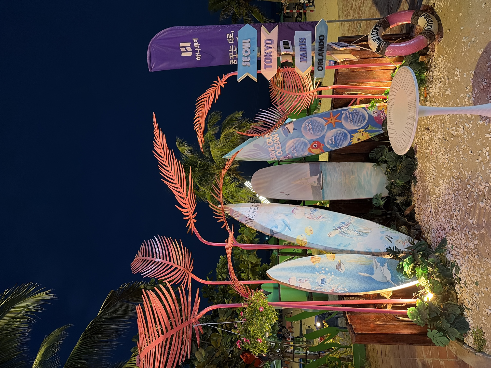 The image depicts an outdoor scene at night, likely a beach resort or themed area in Da Nang, Vietnam. The primary focus is a decorative display featuring several surfboards leaning against a wooden fence and a pink structure resembling palm fronds. The surfboards are adorned with various marine life illustrations, including fish, jellyfish, and turtles, and some have text such as "PINK BEACH" and "SAVE OUR OCEAN."
To the right of the surfboards, a purple flag with Korean text and a stylized "H" logo stands tall. Below the flag is a directional signpost with multiple arrows pointing to different cities: SEOUL, TOKYO, PARIS, and ORLANDO. A pink life preserver ring with "WELCOME Onboard" written on it hangs from the signpost. In the foreground, a white pedestal table with a circular top is placed on a sandy area covered with small white pebbles. Lush green plants and some lighting are visible at the base of the display. Palm trees with green fronds are silhouetted against the dark night sky. The overall ambiance suggests a tropical, summery theme, possibly an attraction or photo opportunity. There are no people clearly visible in the foreground of the image.